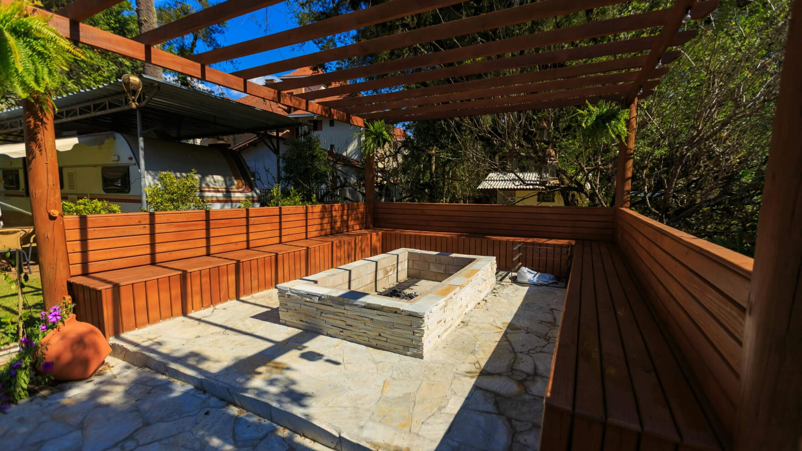 Outdoor wooden pergola with built-in bench seating surrounding a stone fire pit on a stone patio