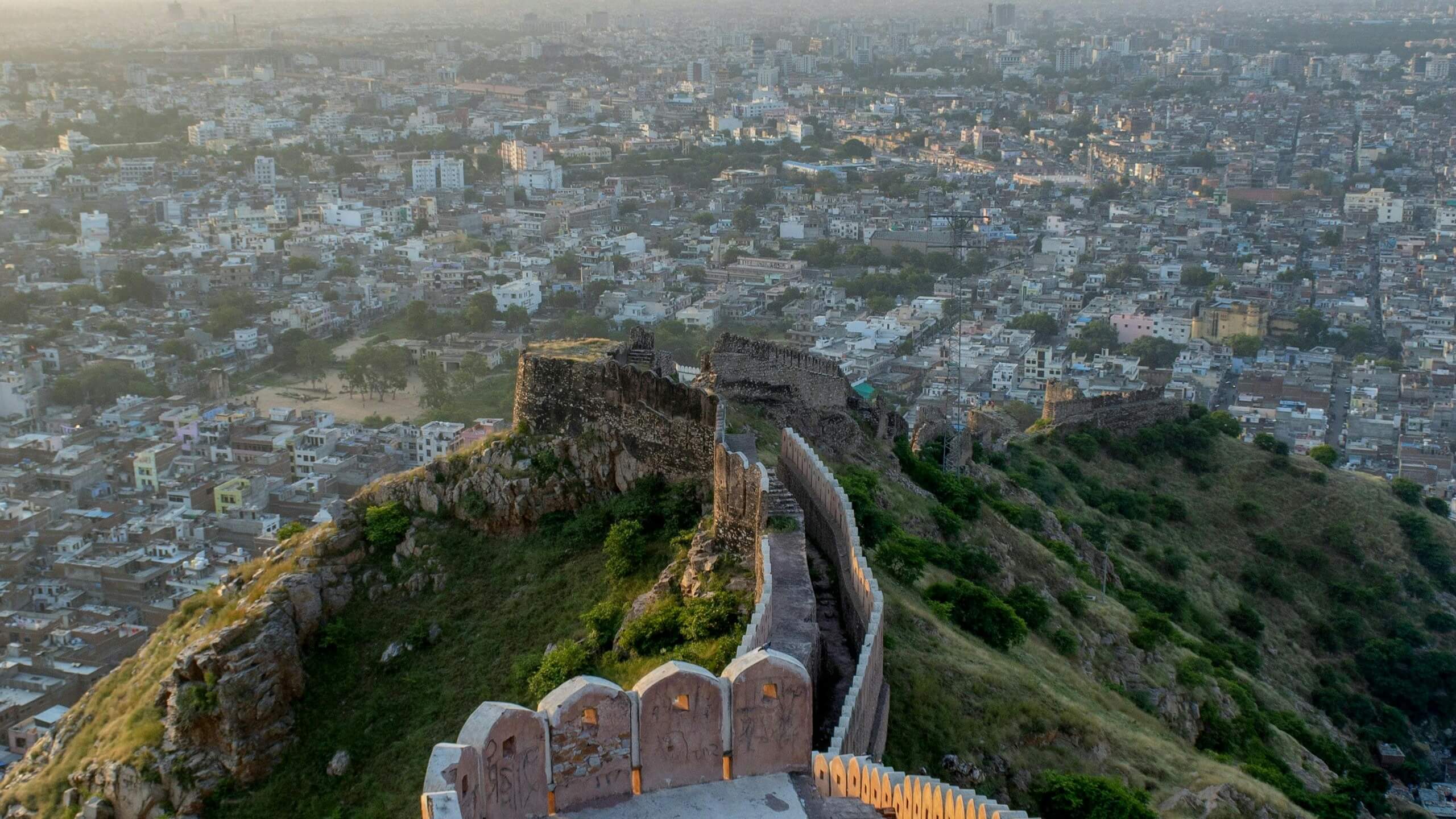 Ancient stone fort wall winding along a green hill overlooking a densely populated cityscape at sunset.