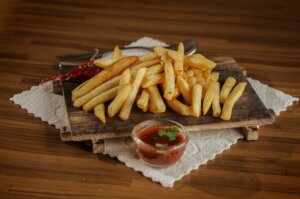 French fries on a wooden board with a small bowl of red dipping sauce and dried red chili peppers on a white cloth.