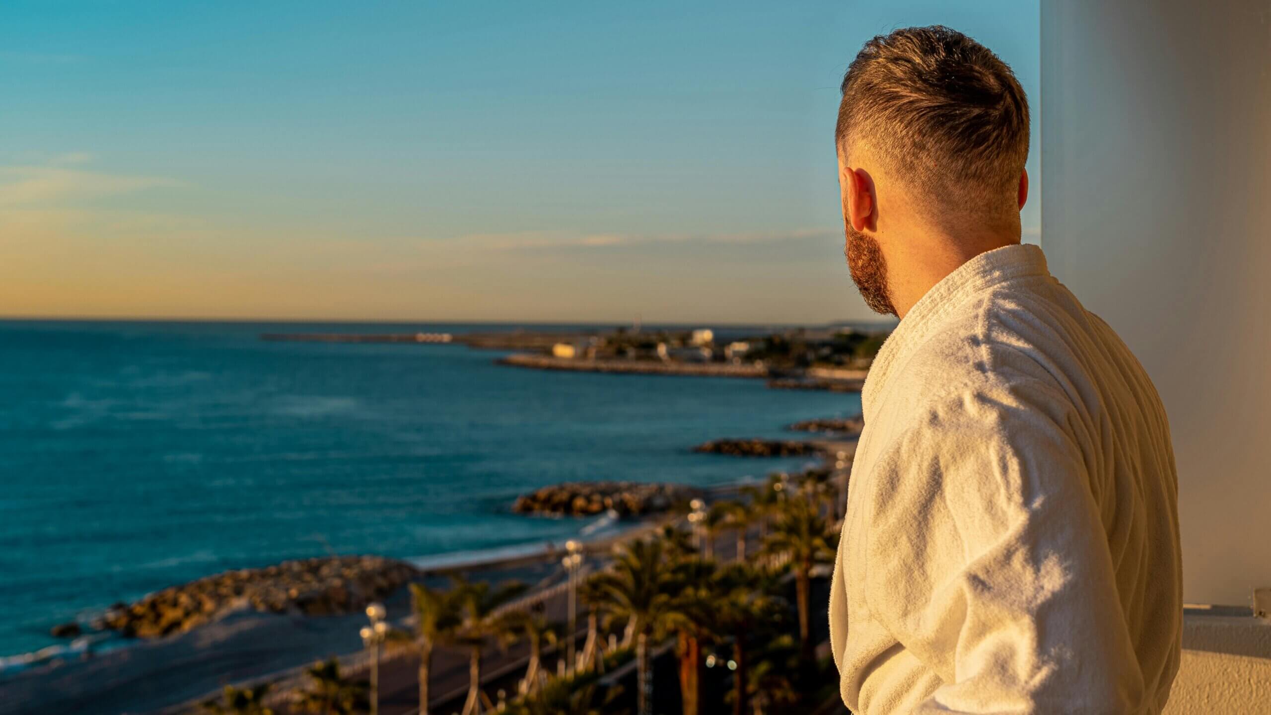 Man in white bathrobe looking at ocean coastline during sunset