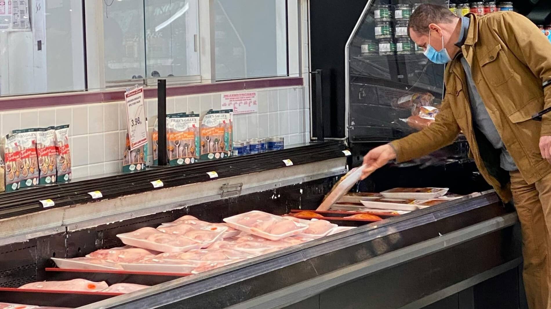 Man wearing a mask and brown jacket selecting packaged chicken from a refrigerated meat display in a grocery store.