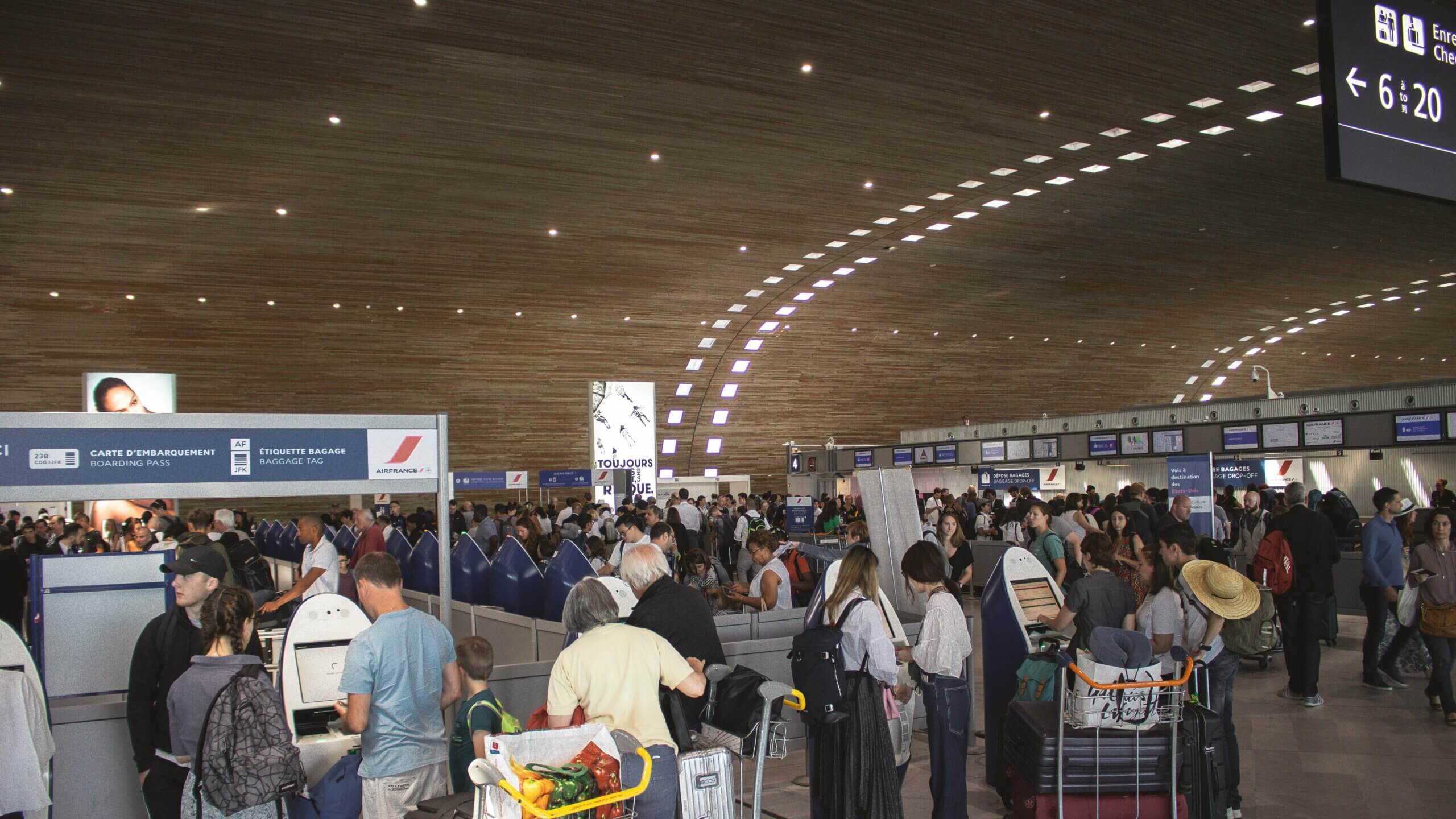 Crowded Air France check-in area at an airport with passengers using self-service kiosks and luggage carts.