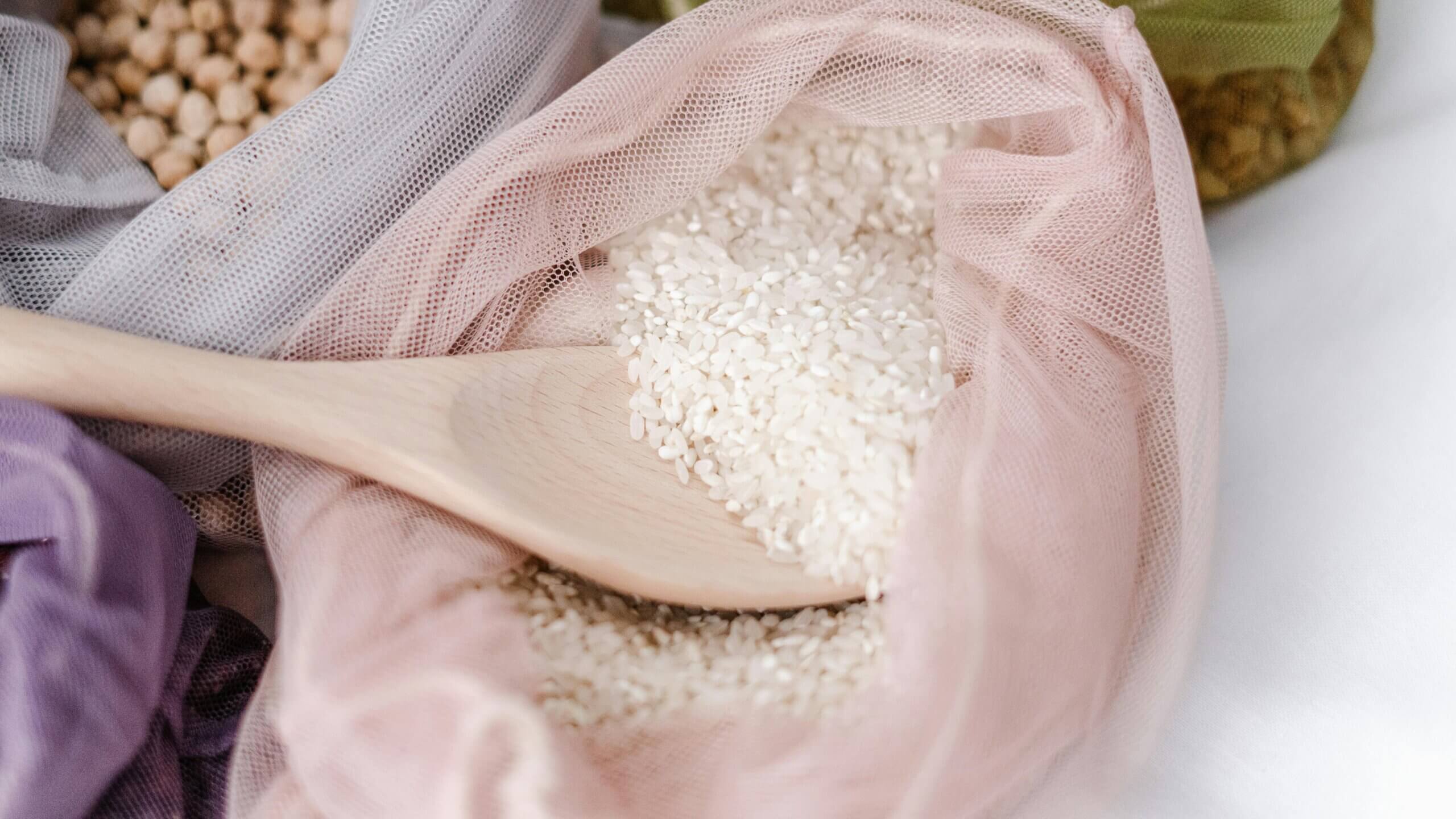 Wooden spoon scooping white rice from a pink mesh produce bag with other legumes in the background