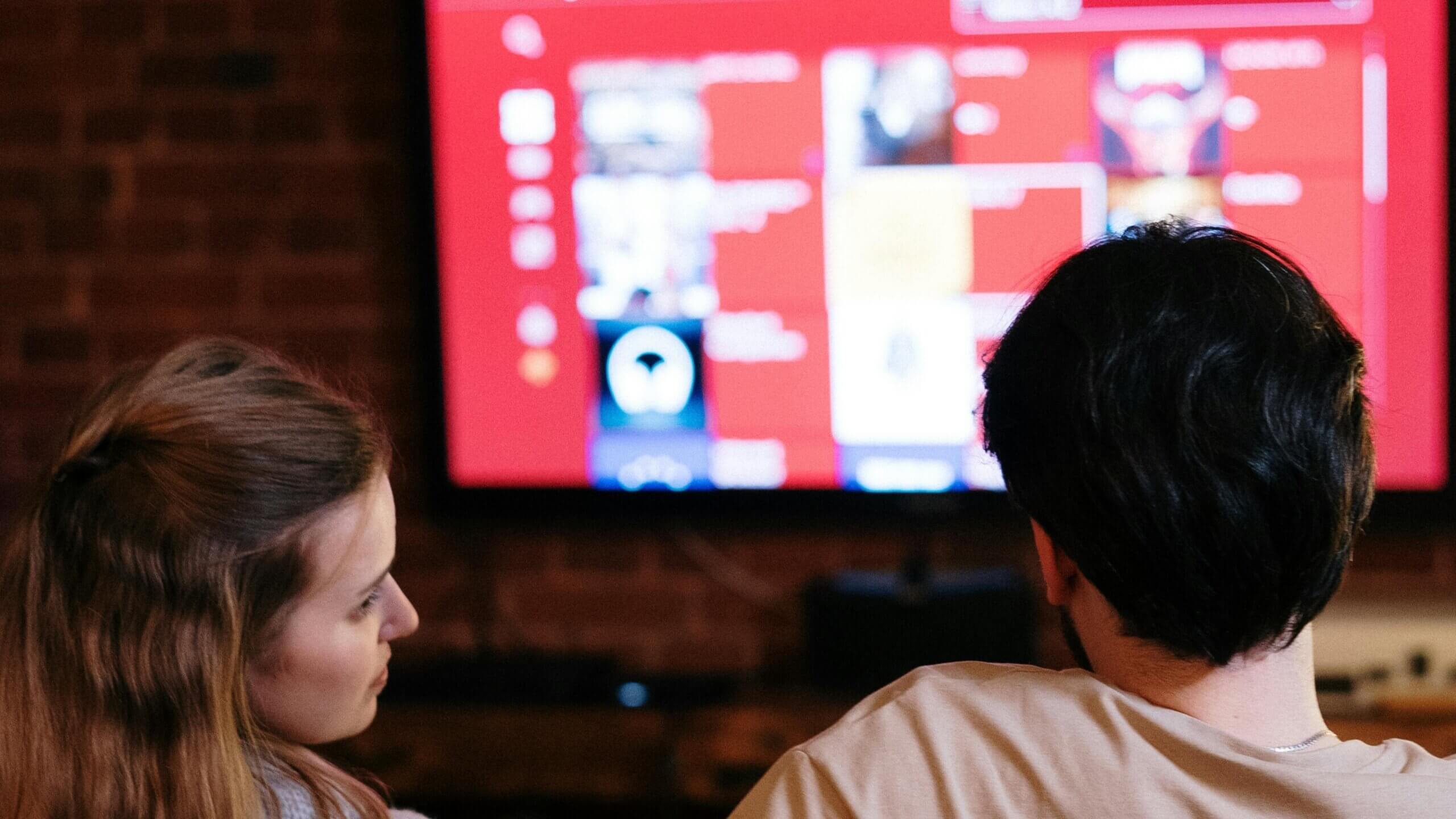 Two people watching a TV screen with a red interface in a dimly lit room