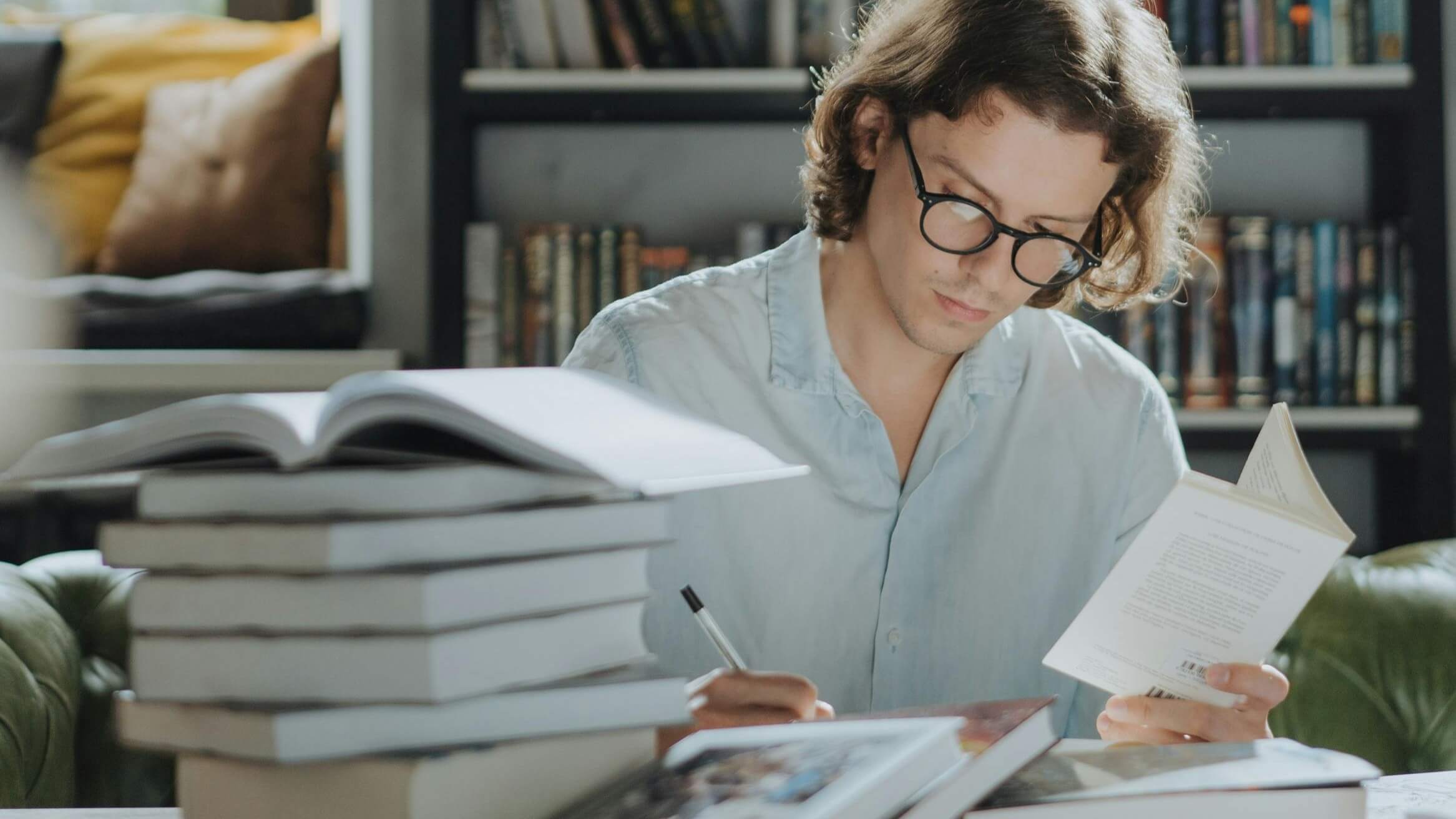 Young man with glasses reading and taking notes surrounded by books in a library setting