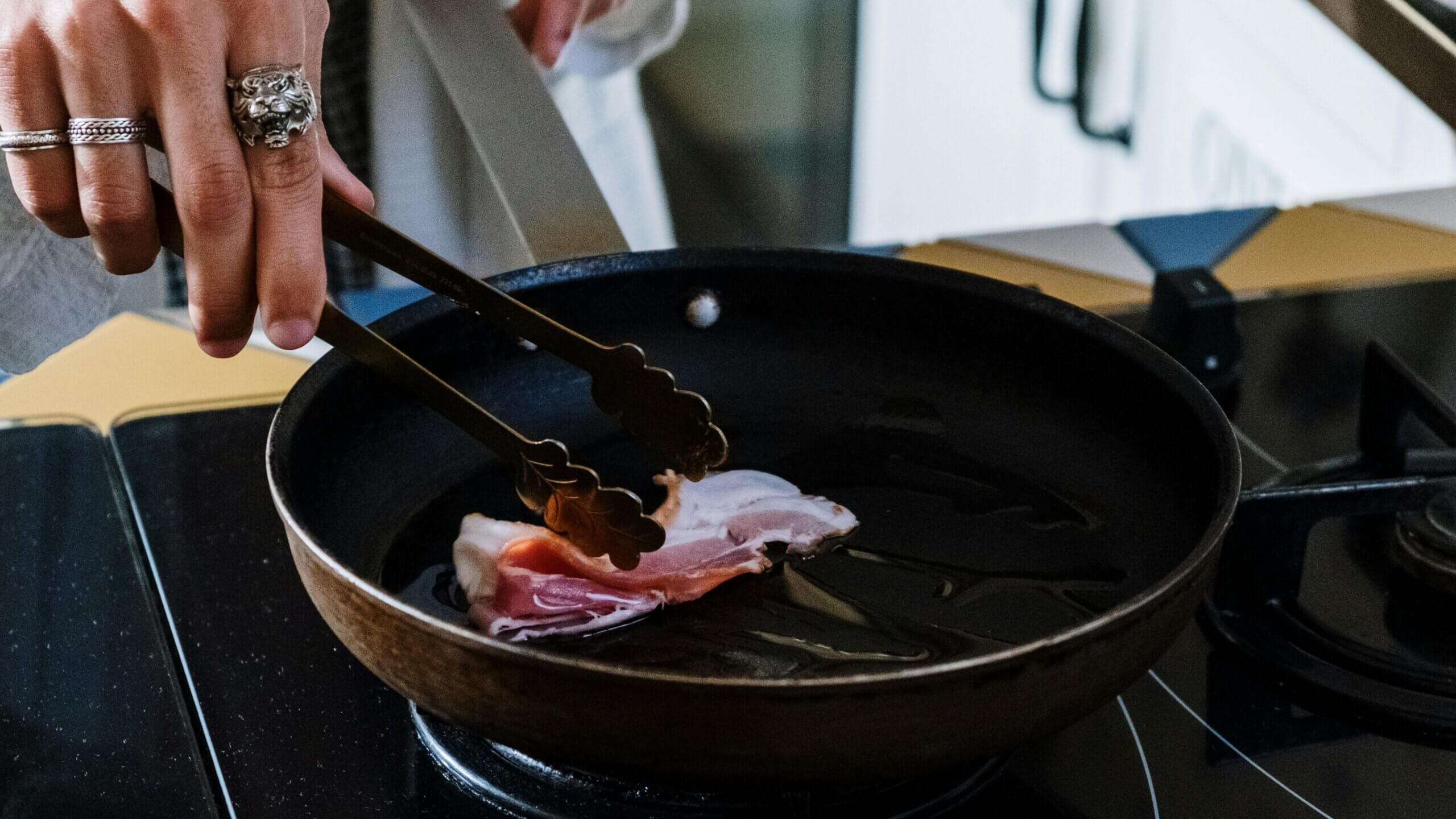 Hand with rings using tongs to cook bacon in a frying pan on a stovetop.