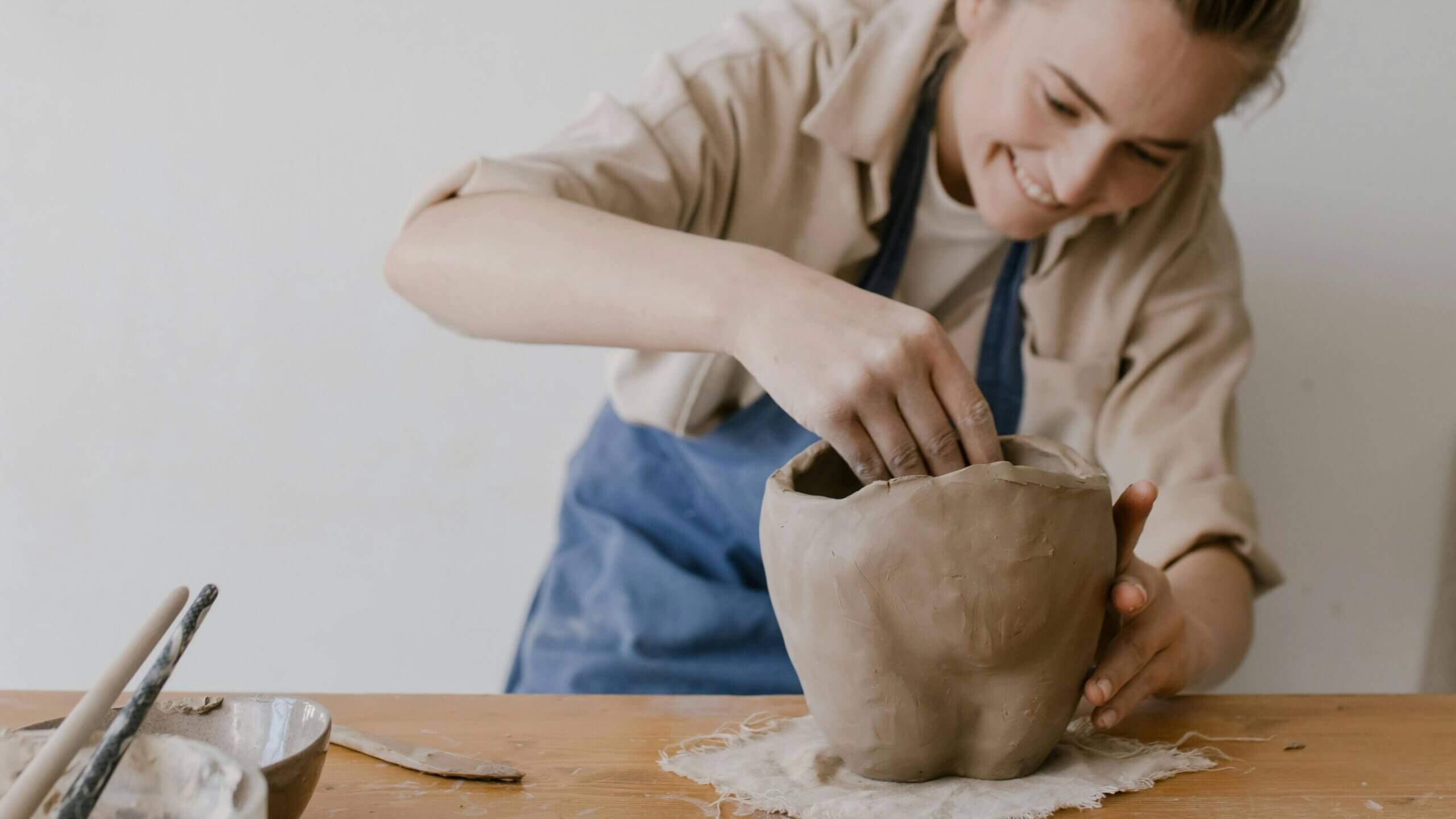 Person shaping a large clay pot on a wooden table while smiling and wearing a blue apron.