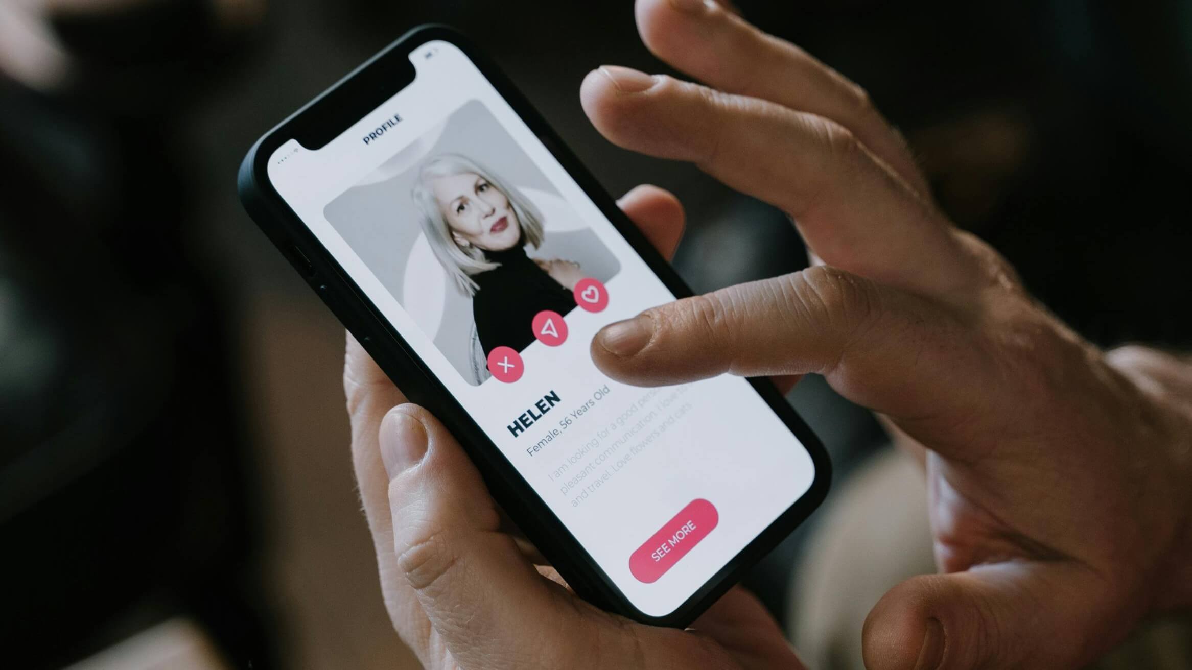 Person holding smartphone showing dating profile of Helen, 56, with photo and interaction buttons.