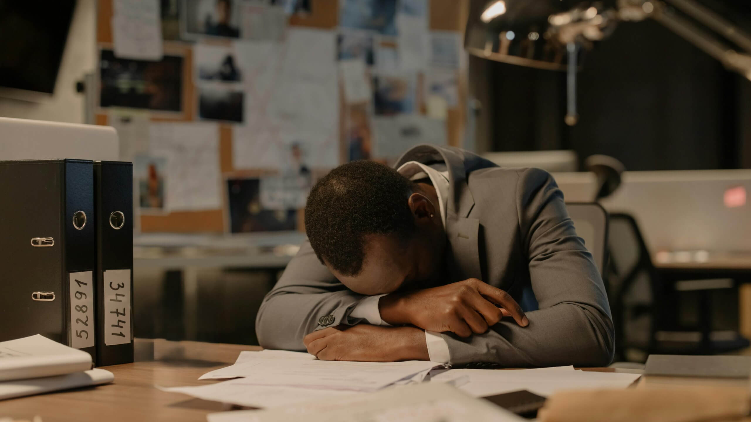 Man in gray suit resting head on crossed arms at cluttered office desk with binders and papers