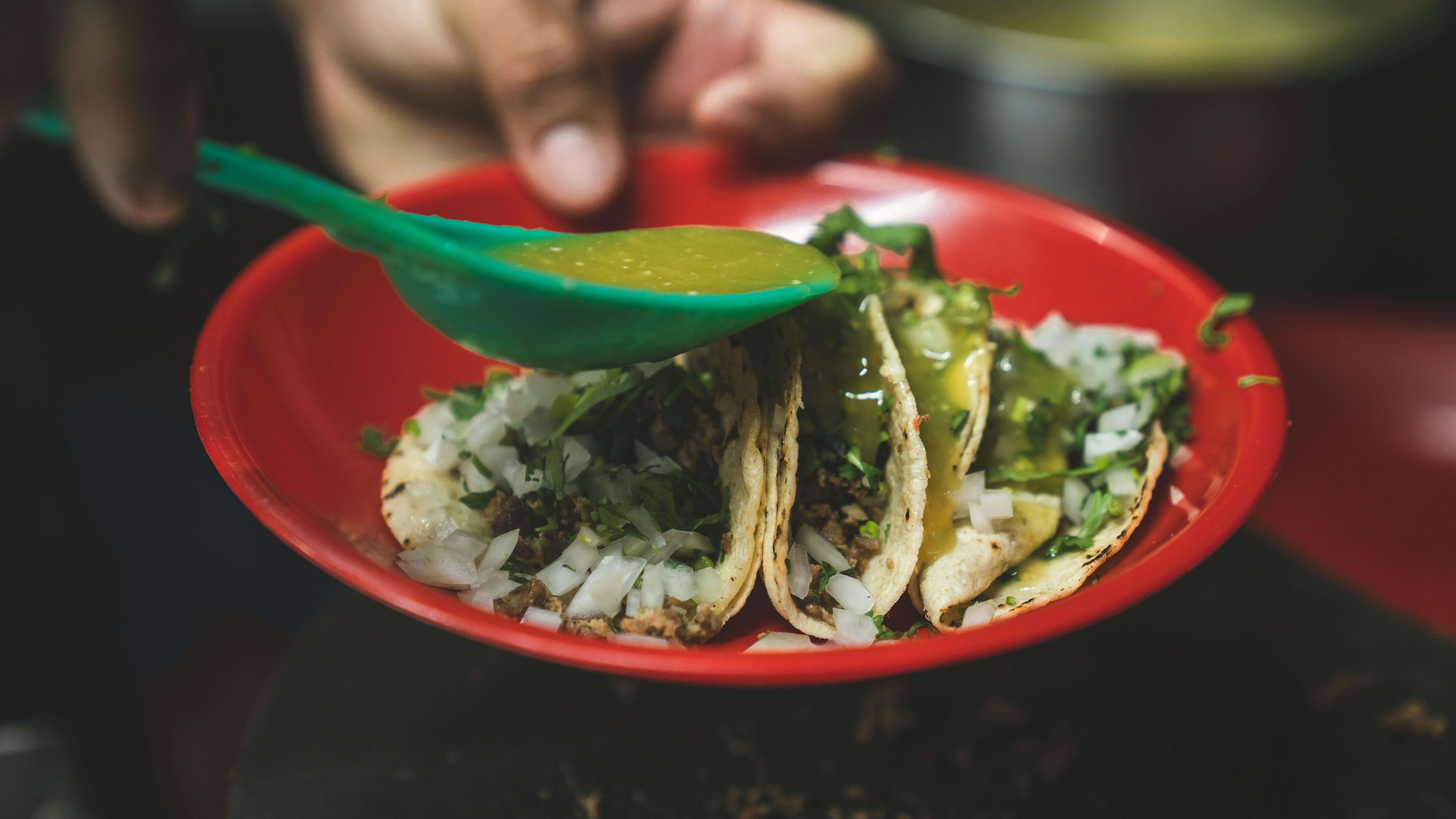 Three tacos with chopped onions and cilantro on a red plate being topped with green salsa from a green spoon