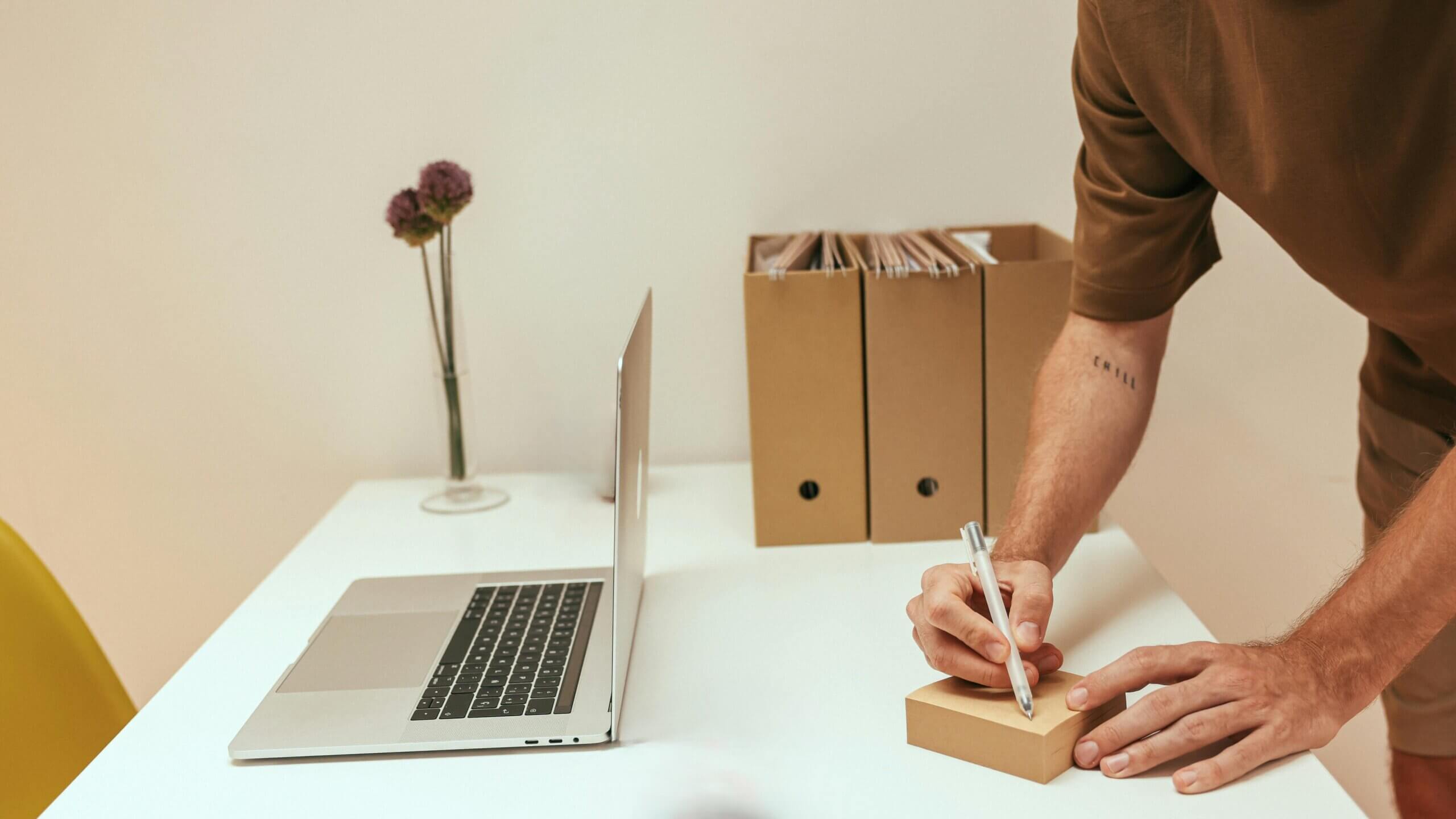 Person writing on a small brown box on a white desk with a laptop, file folders, and a vase with flowers nearby