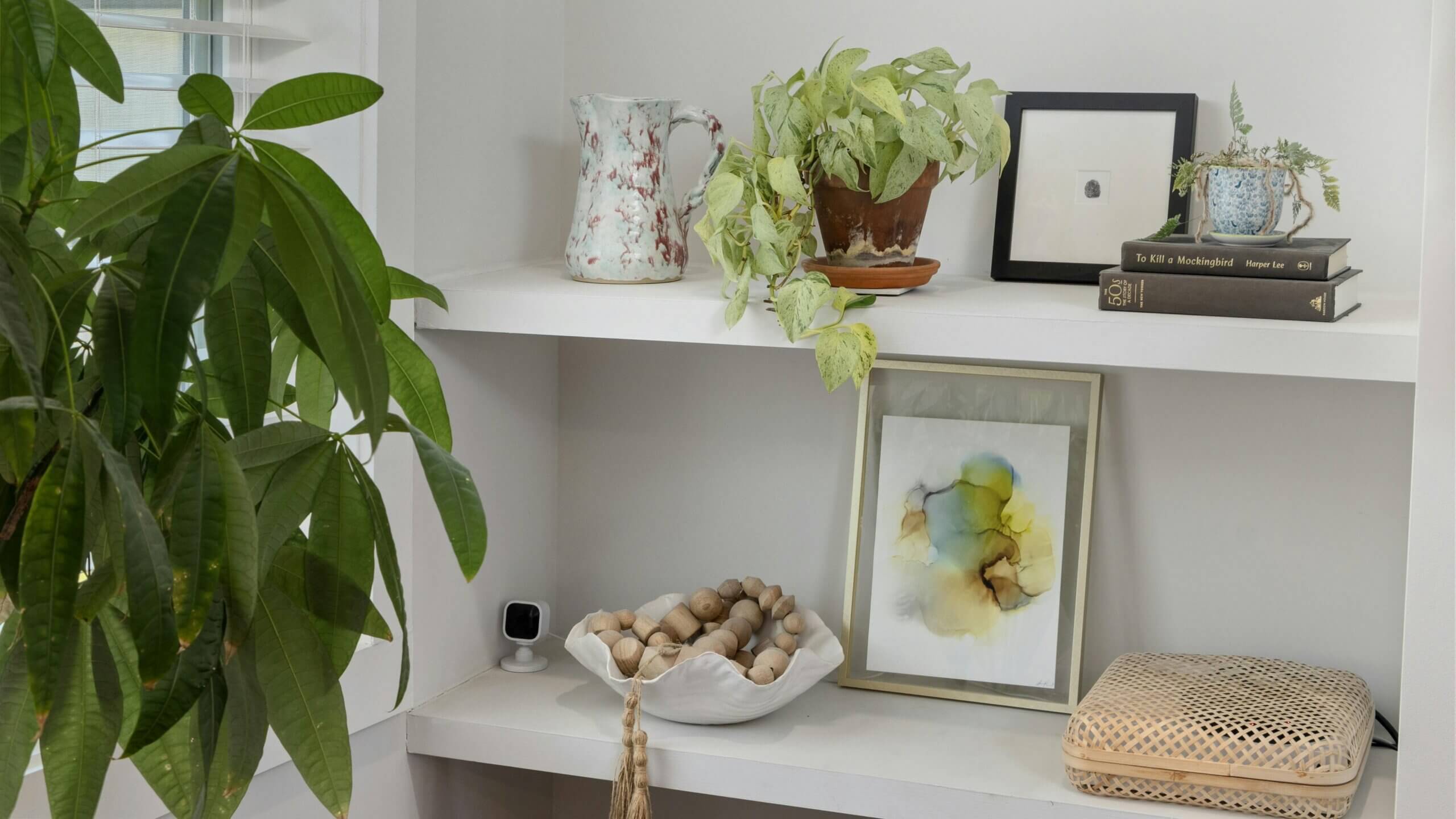 White shelves with potted plants, a ceramic pitcher, framed art, books including "To Kill a Mockingbird," and decorative wooden beads.