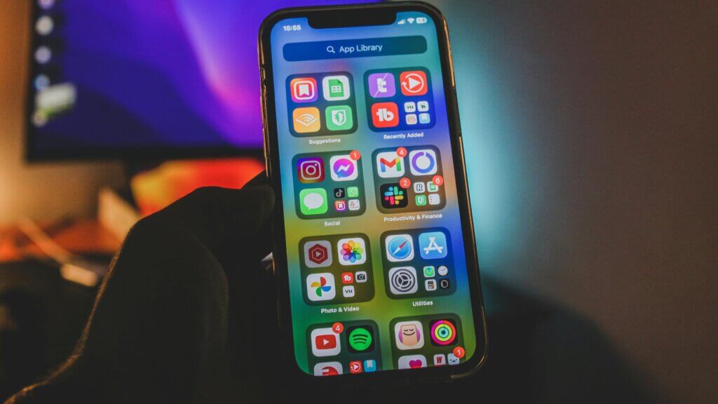 Hand holding iPhone displaying App Library with social, productivity, photo, and utility app folders visible