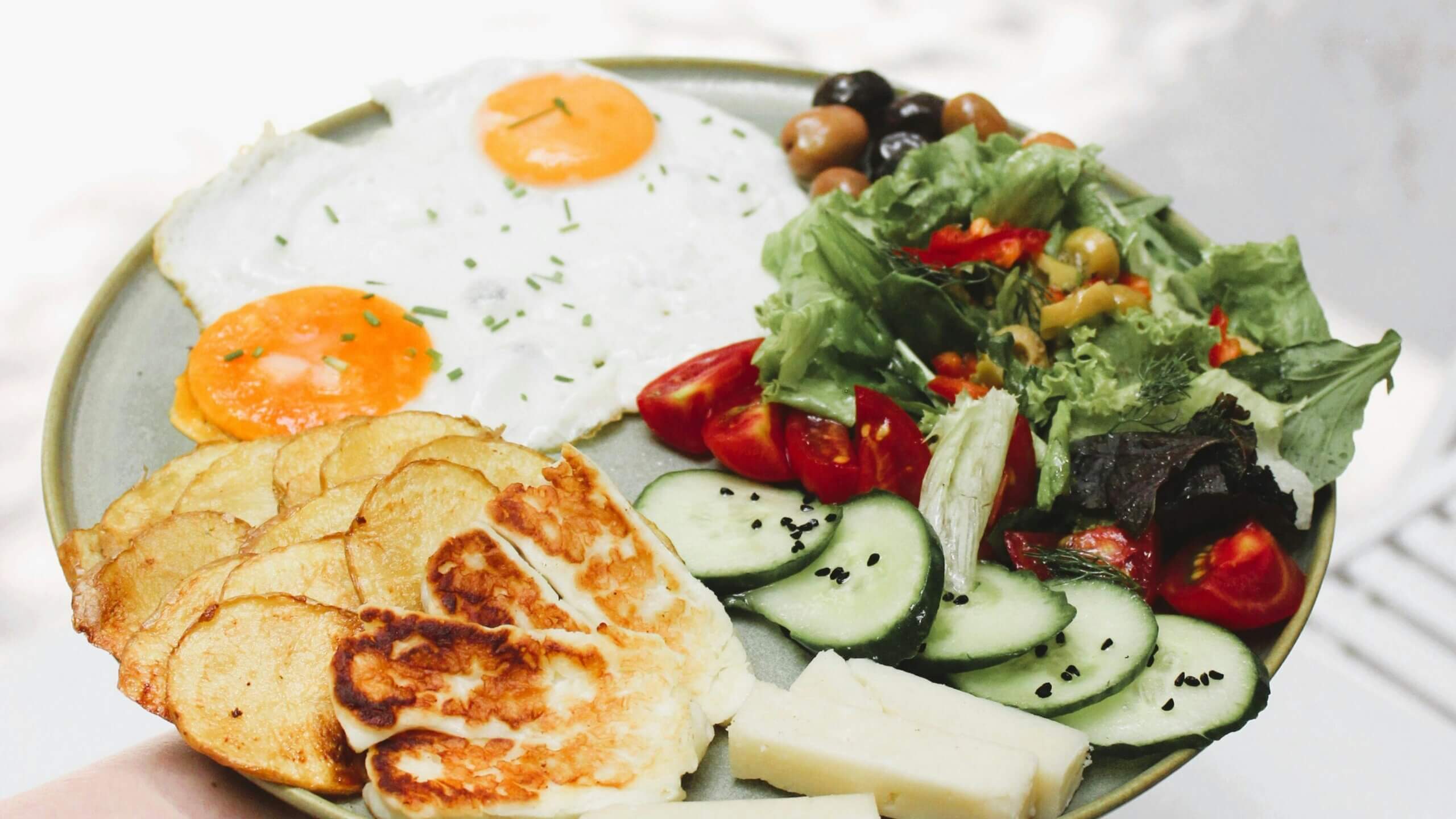 Plate with fried eggs, sliced potatoes, grilled halloumi, cucumber, tomato, olives, and mixed salad greens