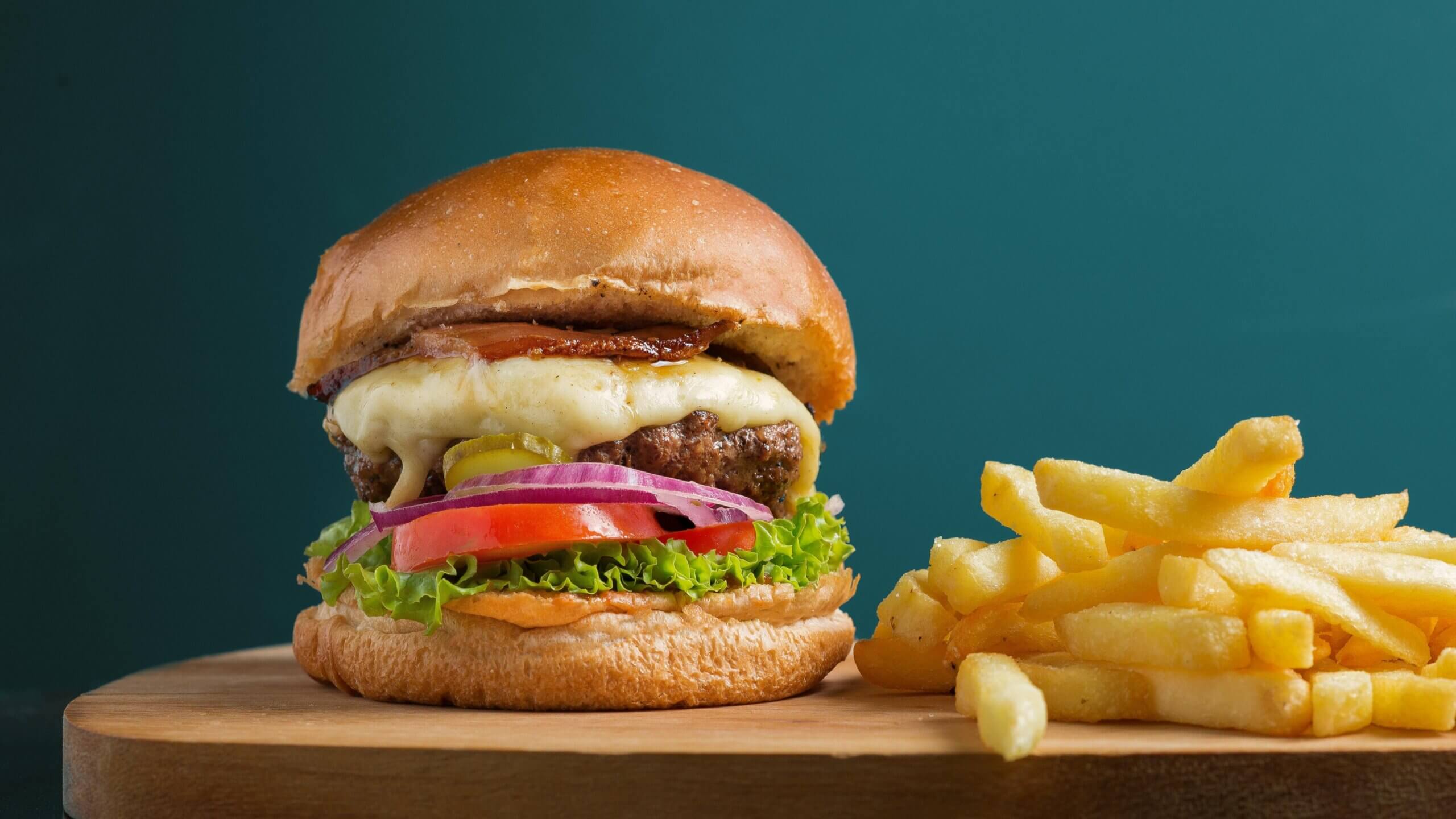 Cheeseburger with lettuce, tomato, onion, pickles, bacon, and a side of French fries on a wooden board