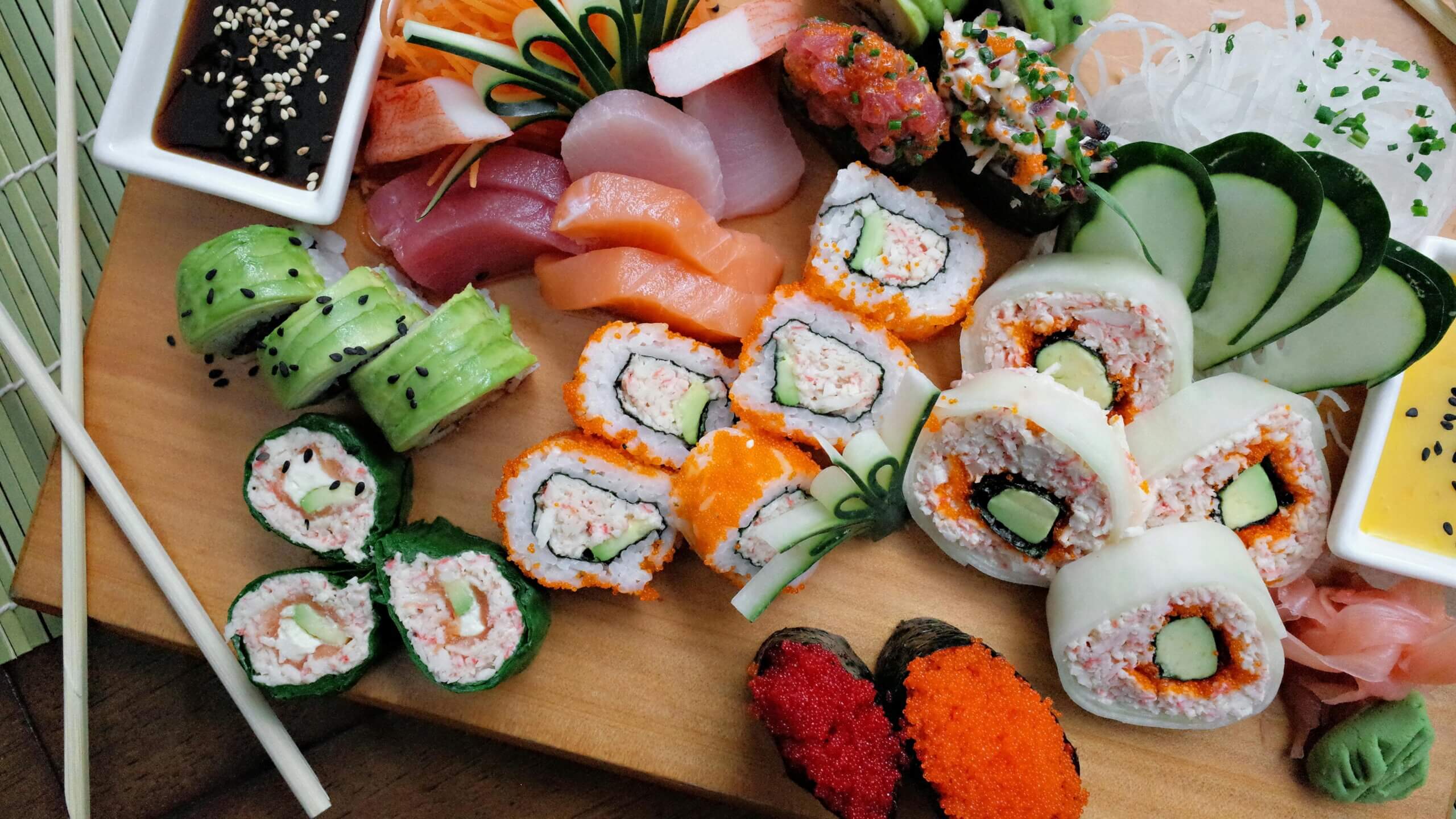 Assorted sushi rolls and sashimi with cucumber, wasabi, ginger, and dipping sauces on a wooden board.