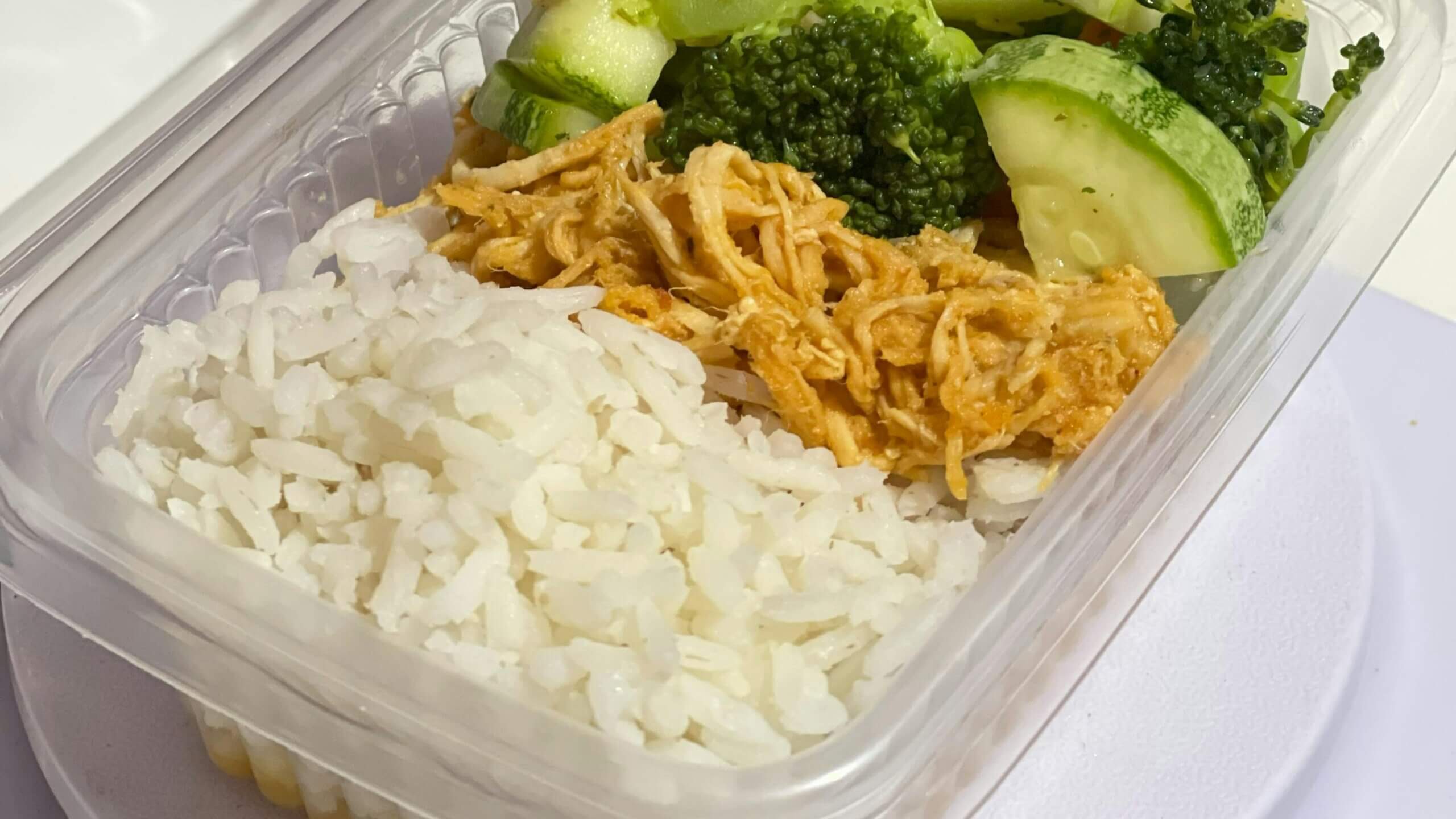 Takeout container with white rice, shredded chicken, broccoli, and cucumber slices