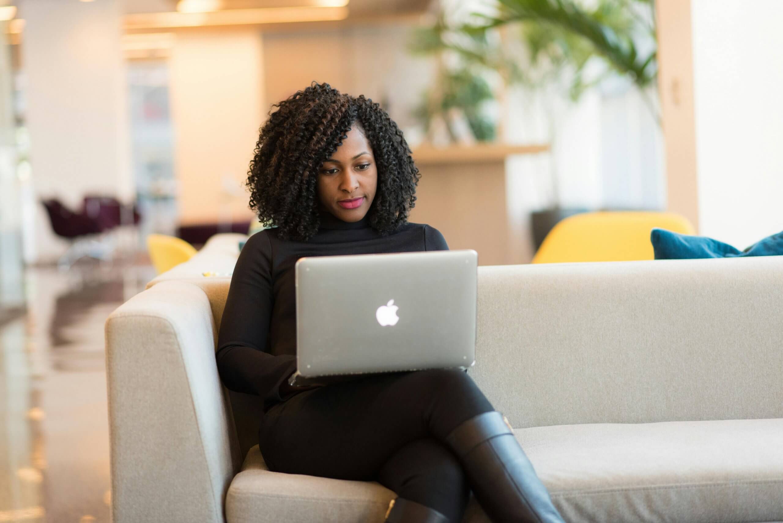 Woman with curly hair sitting on a beige couch using a silver Apple MacBook laptop in a modern office lounge