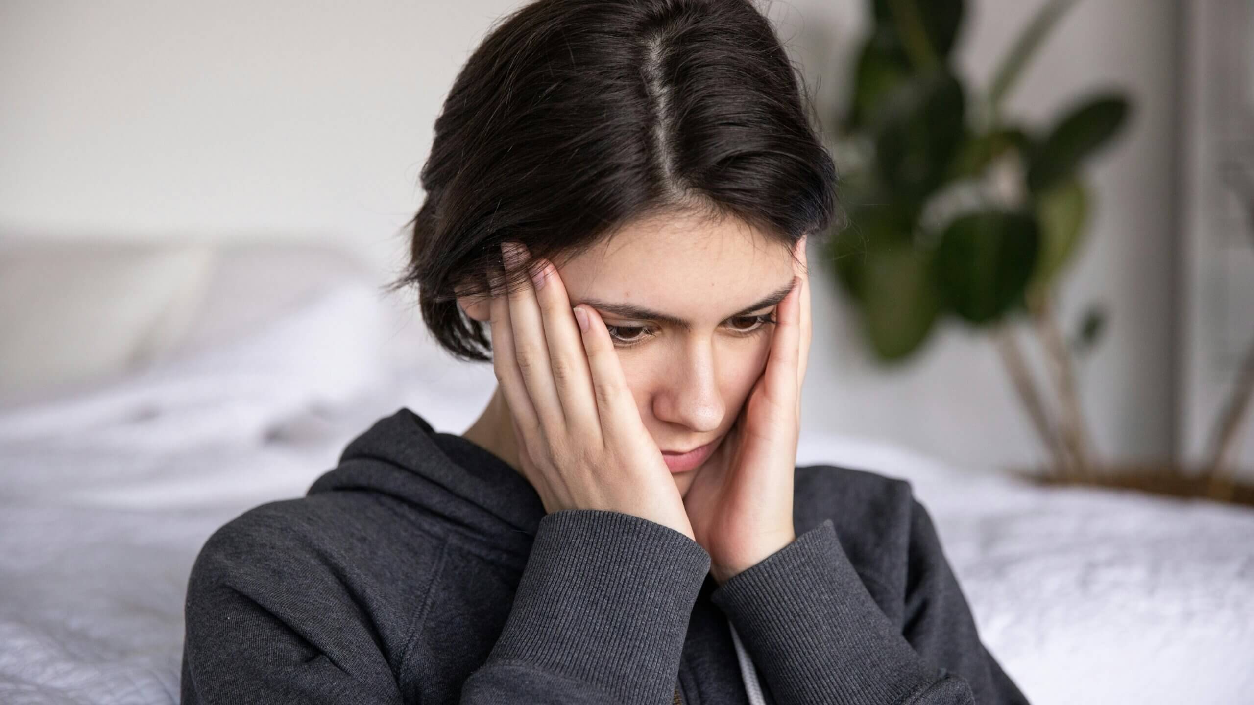 Young woman in a dark hoodie holding her face, appearing worried or stressed indoors.
