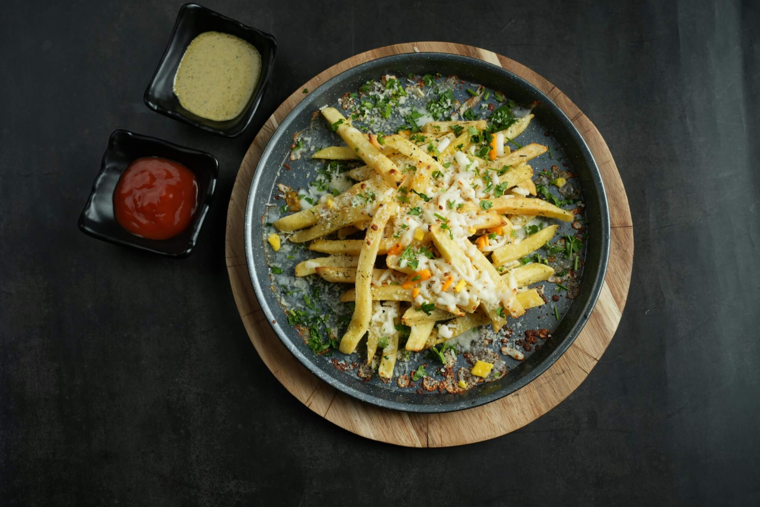 Cheese-covered French fries garnished with herbs on a round tray with ketchup and mustard sauces.