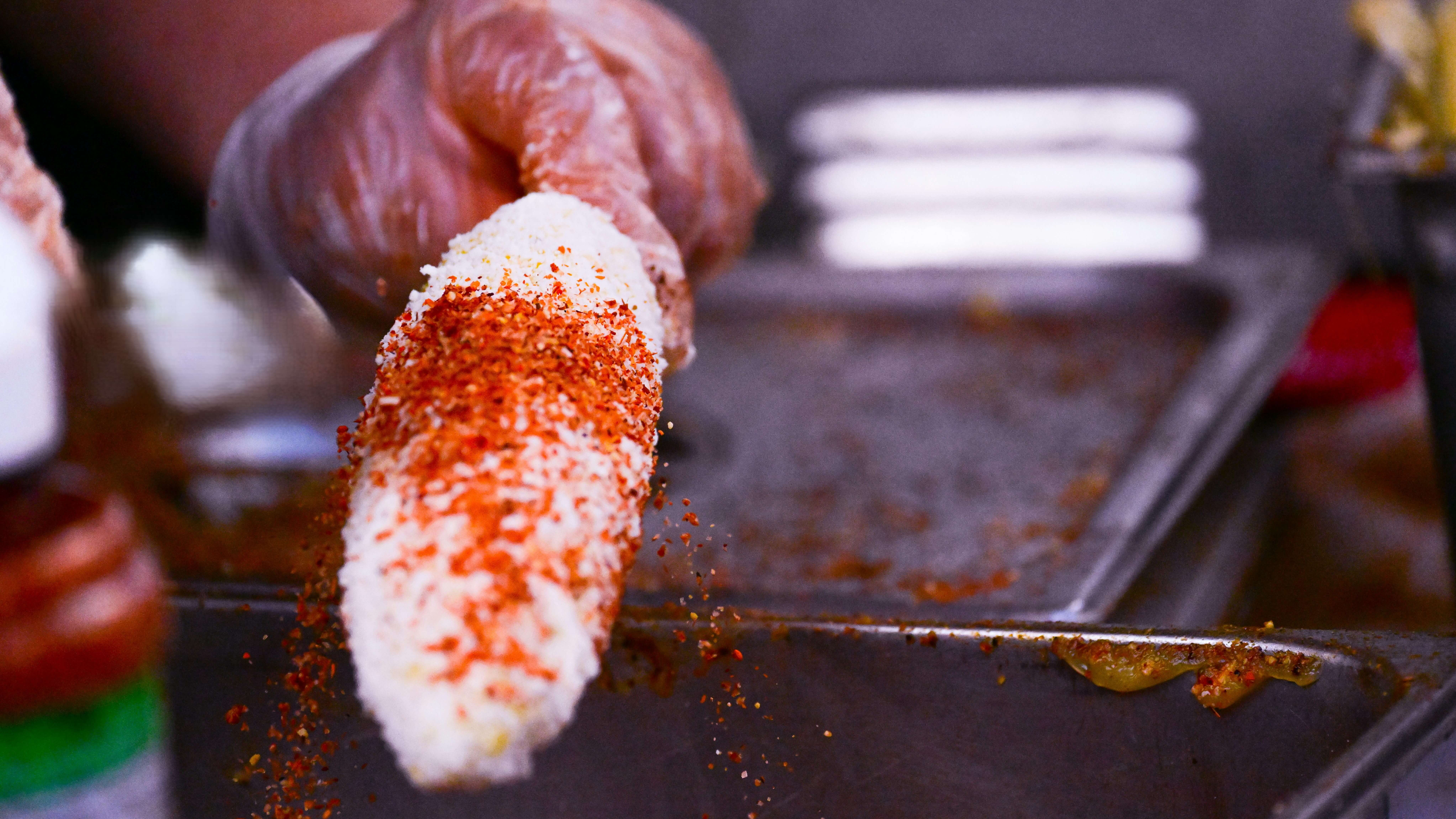 Hand wearing glove sprinkling chili powder on Mexican street corn (elote) with cheese coating.
