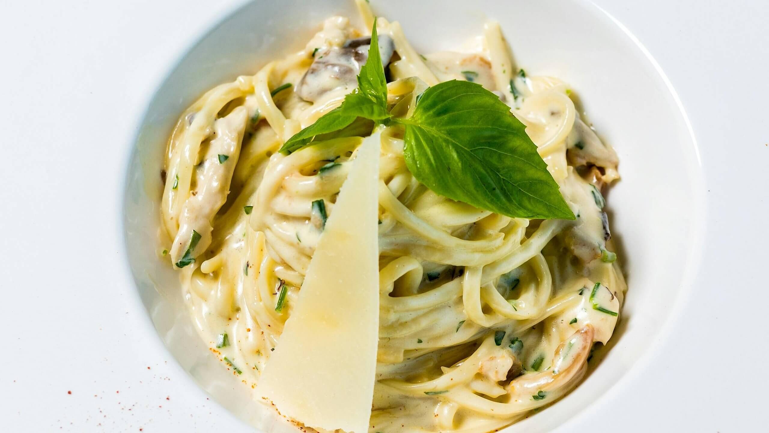 Creamy fettuccine pasta with herbs, mushrooms, a basil leaf, and a slice of Parmesan cheese in a white bowl