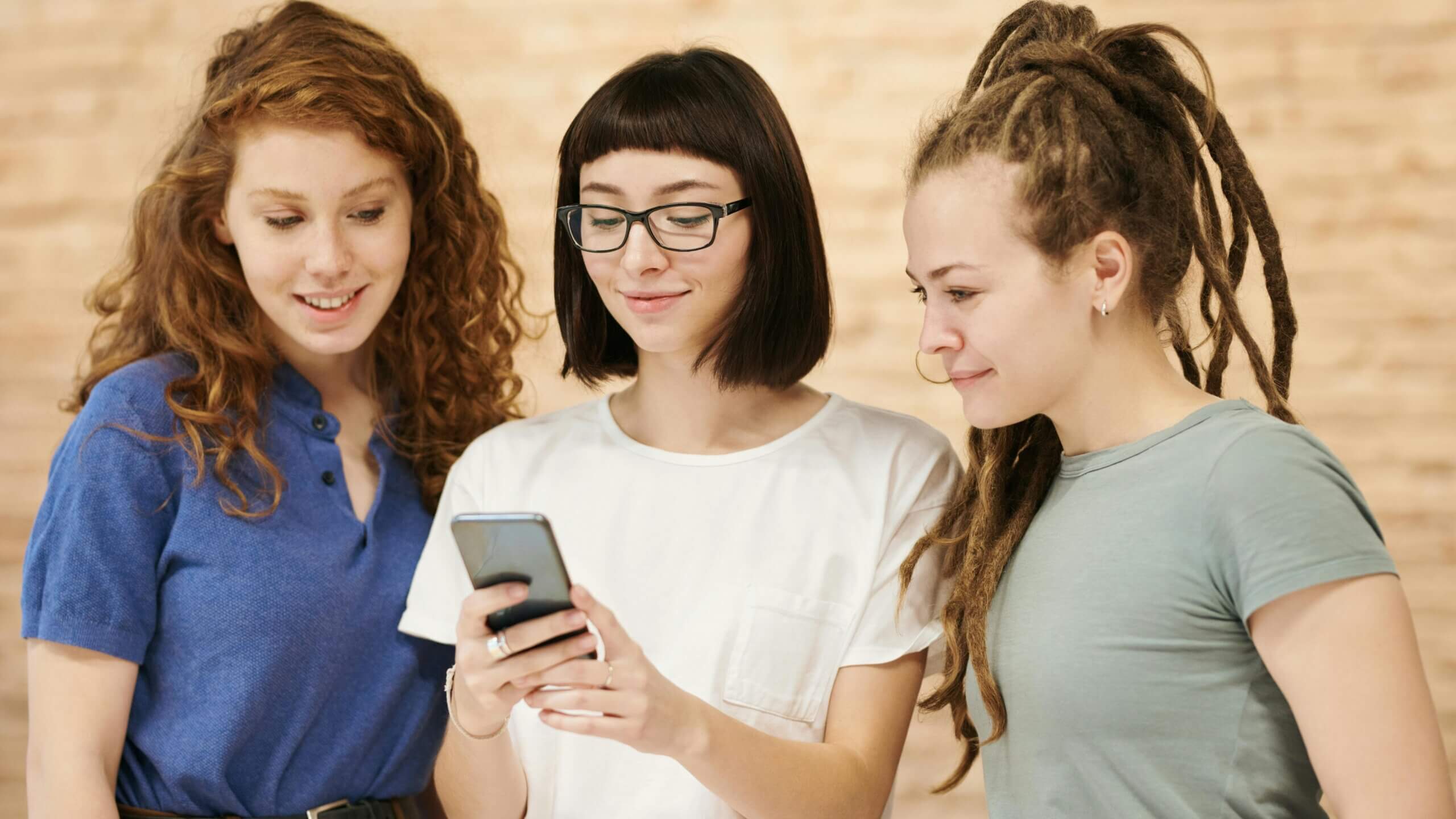 Three young women looking at a smartphone, one with glasses and a white shirt, another with curly red hair in a blue shirt, and one with dreadlocks in a green shirt.