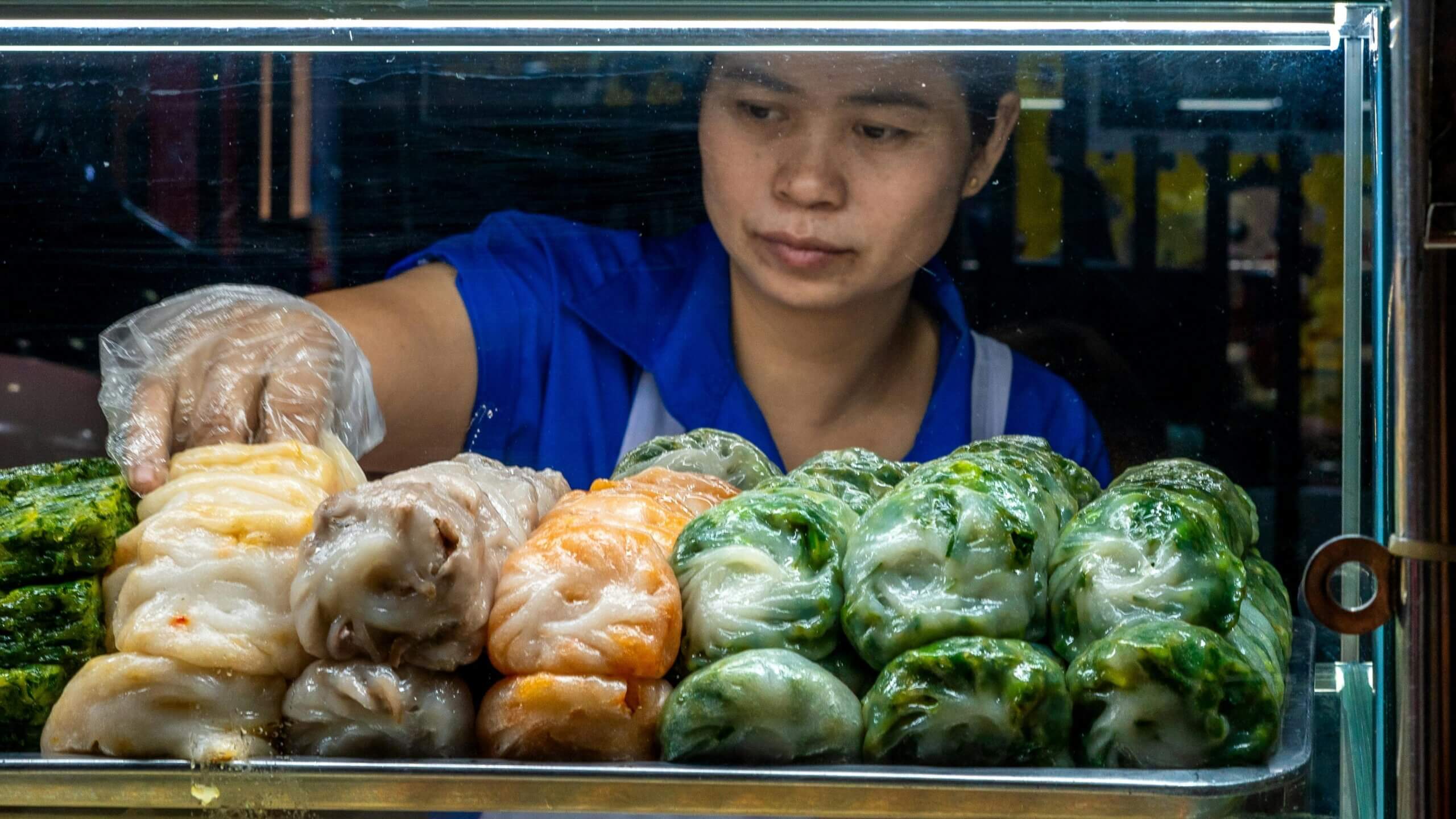 Vendor wearing blue shirt and plastic glove arranging colorful steamed dumplings in a glass display case.