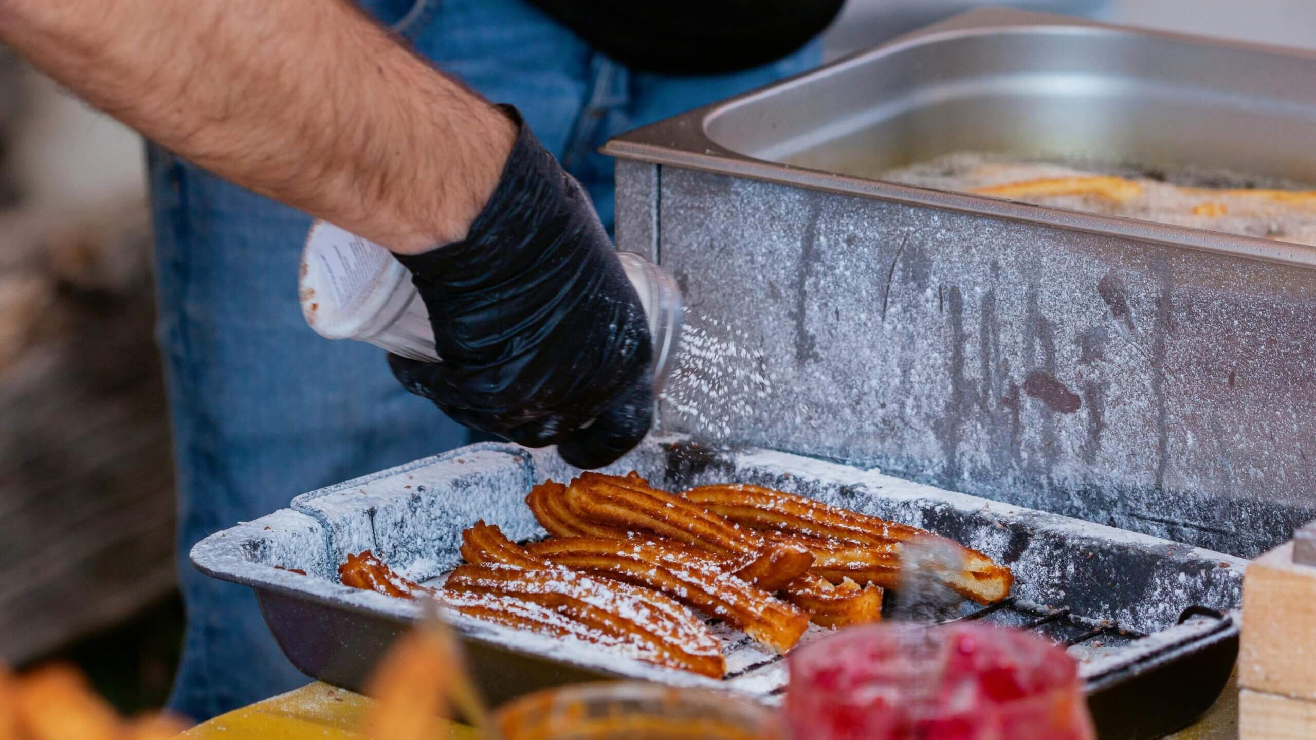 Hand wearing black glove sprinkling powdered sugar on fried churros in a metal tray next to a fryer.