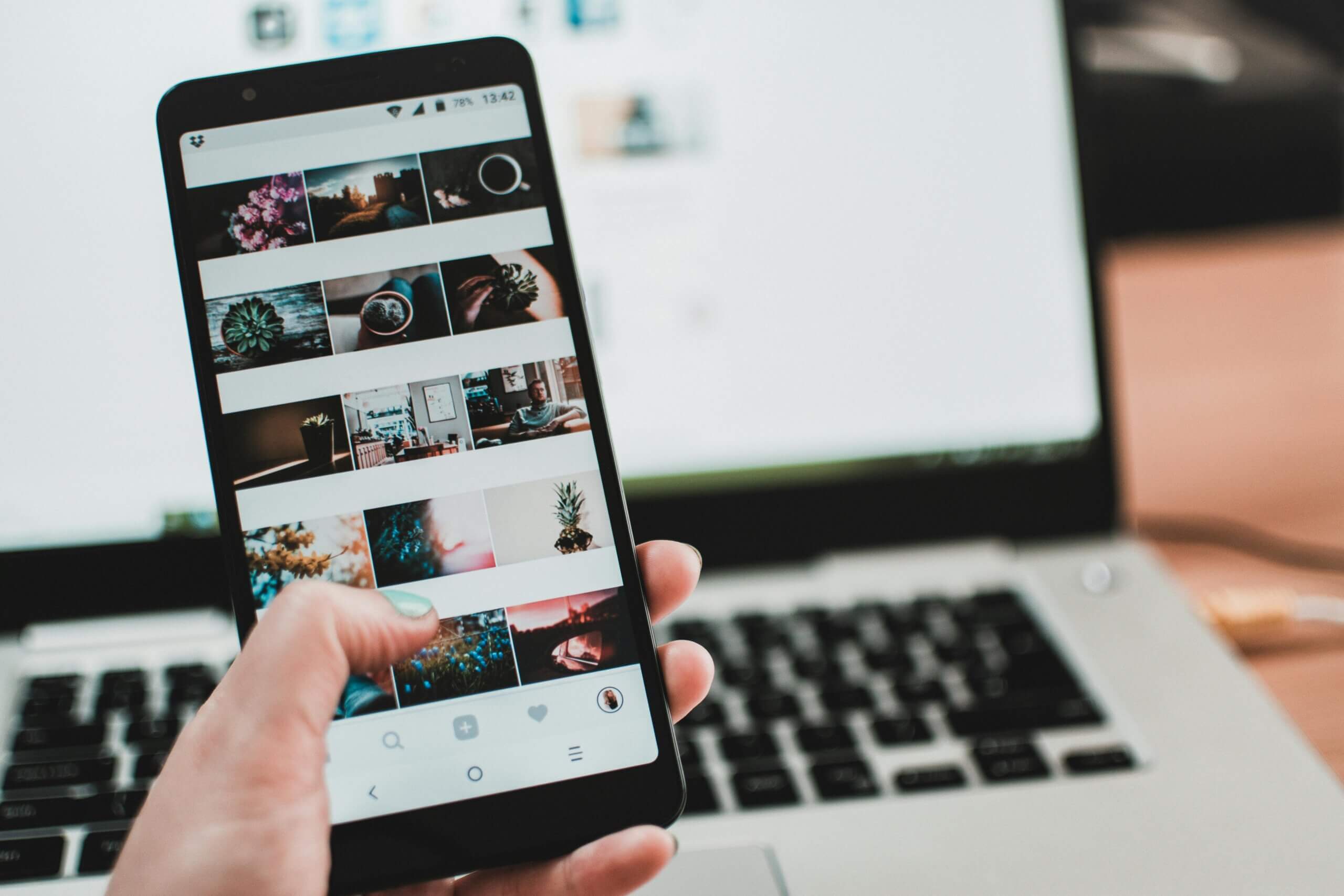 Hand holding smartphone displaying photo gallery app with various images, laptop keyboard in background.