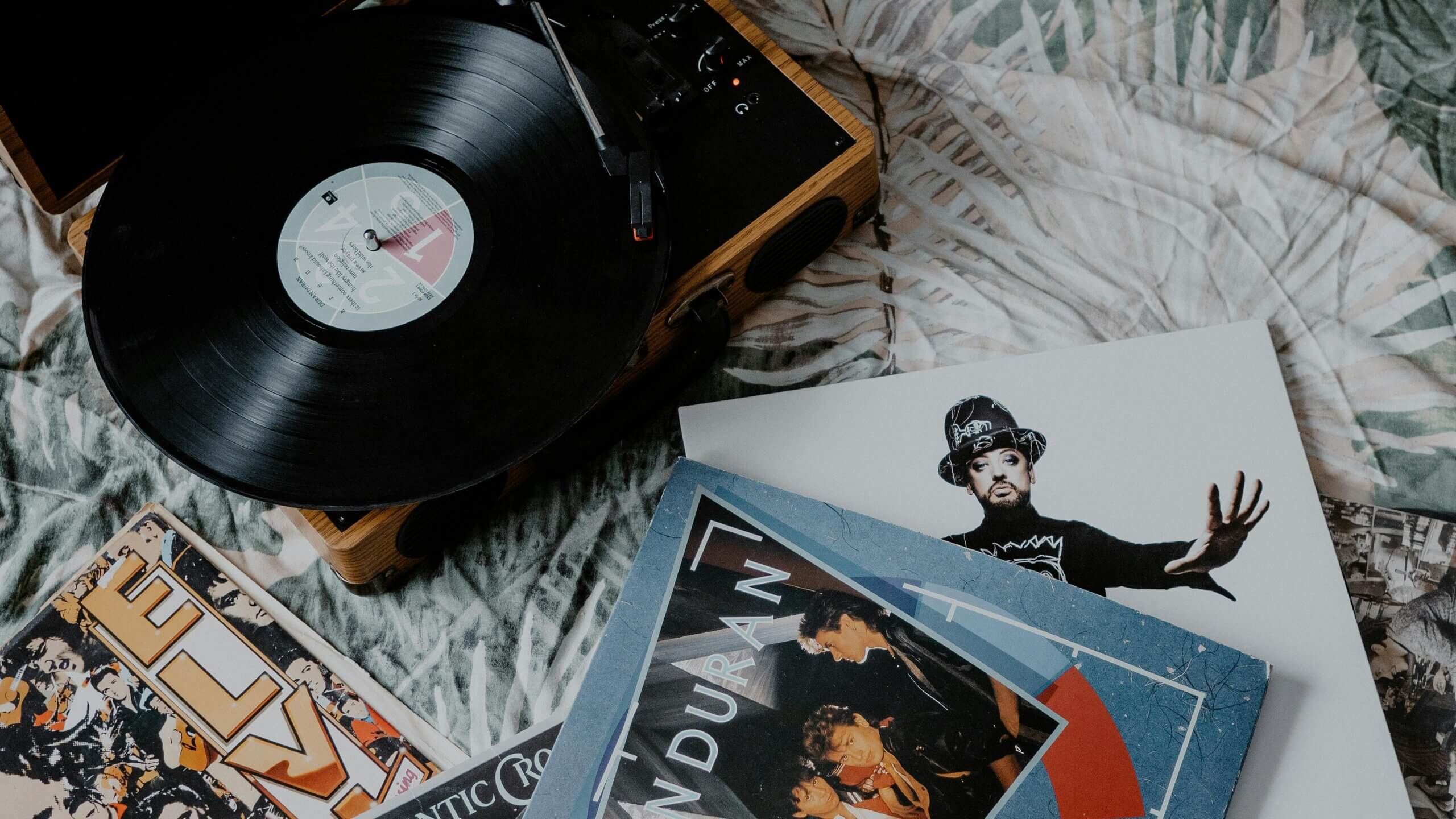 Vinyl record playing on a wooden turntable surrounded by albums including Duran Duran and a man in a hat on a cover.