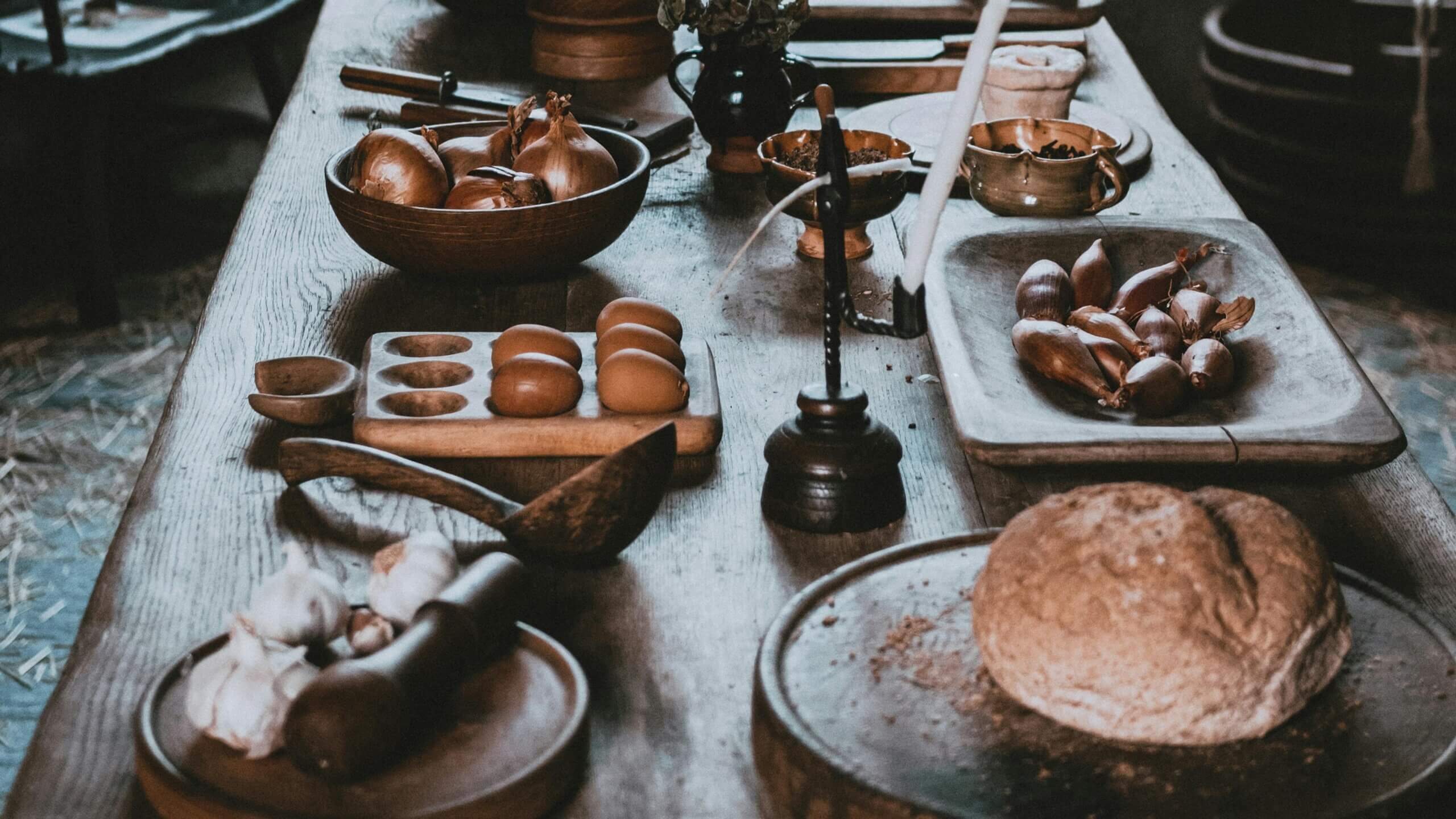 Rustic wooden table with bread, garlic, onions, eggs, and kitchen utensils arranged on it.