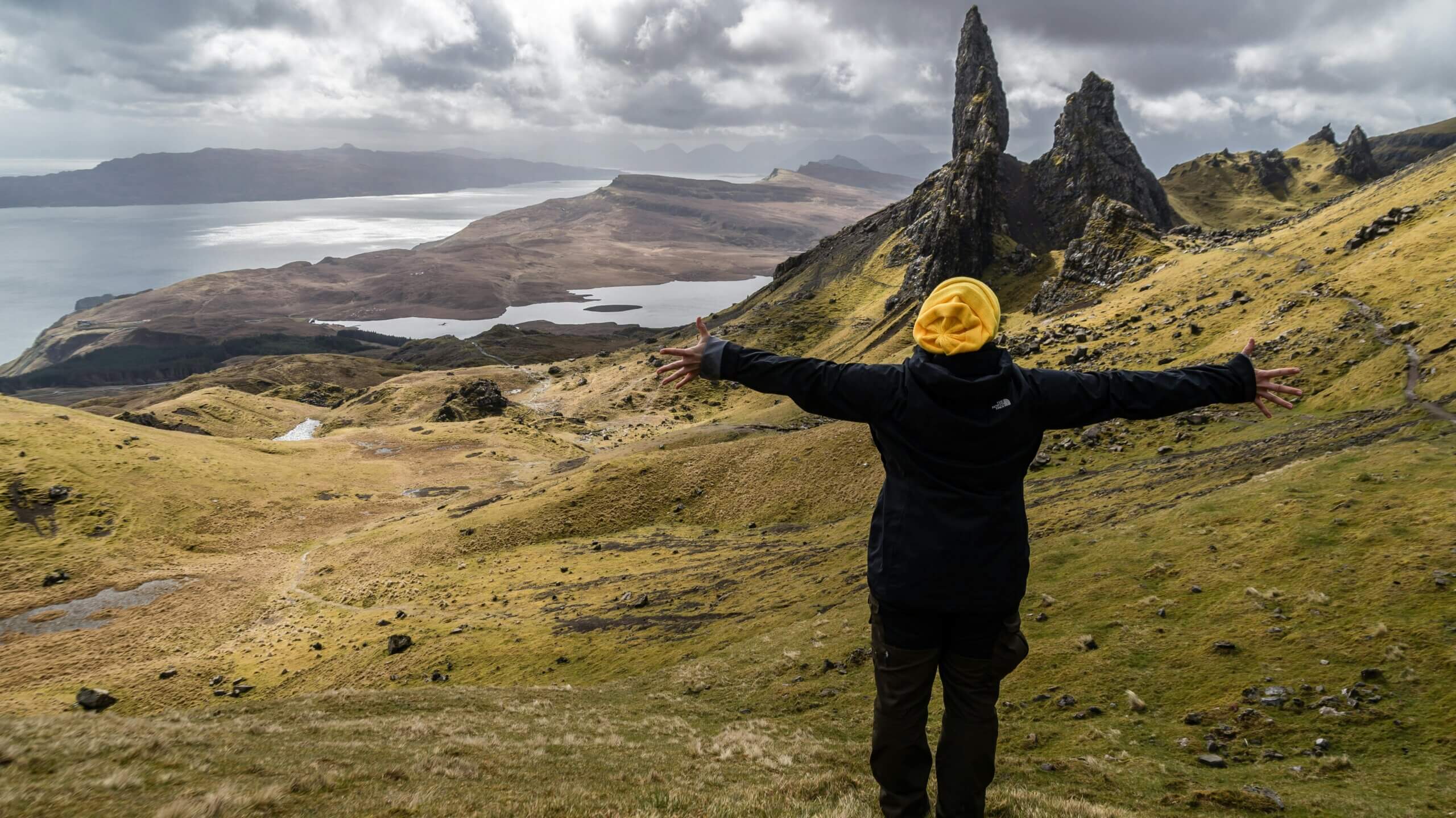 Person in a yellow beanie and black North Face jacket with arms outstretched overlooking the Old Man of Storr rock formation in Scotland