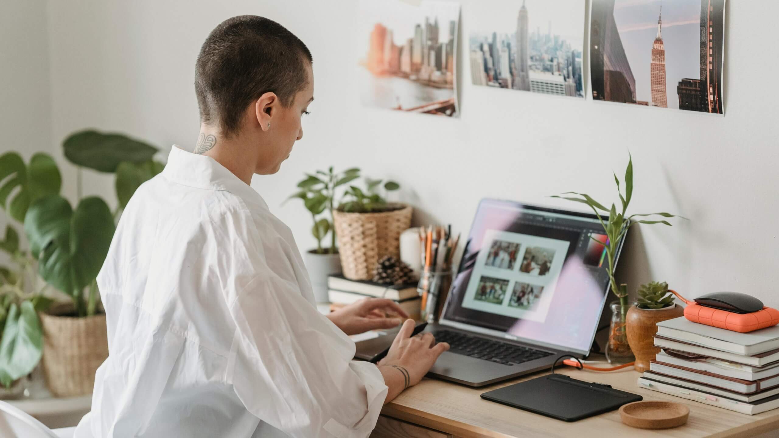 Person with short hair and white shirt working on a laptop at a desk with plants and photos of cityscapes on the wall
