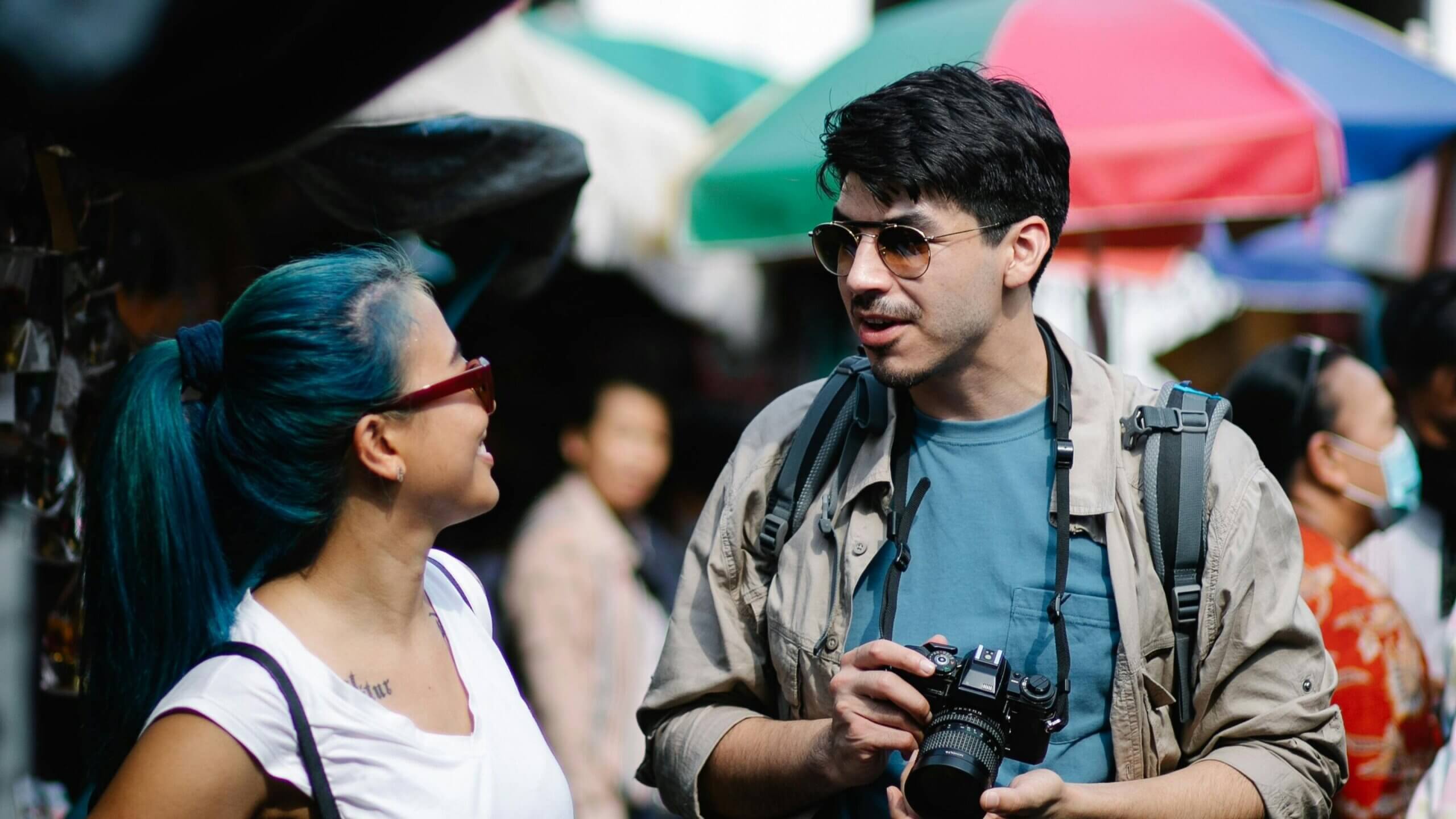Man holding a camera talking to a woman with blue hair and sunglasses at an outdoor market with colorful umbrellas