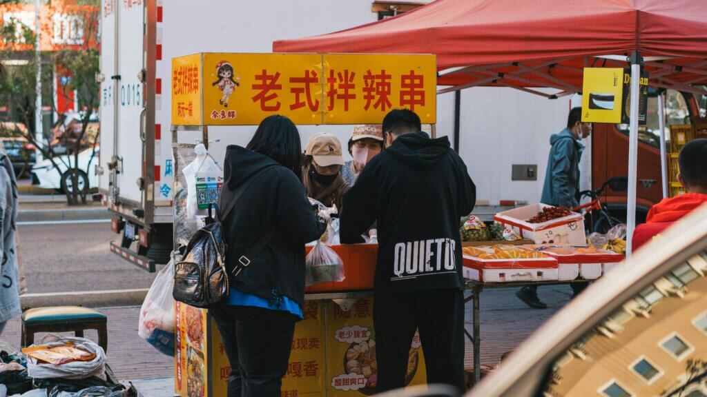 People buying food at a street vendor stall with a yellow sign in Chinese characters under a red canopy