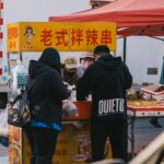 People buying food at a street vendor stall with a yellow sign in Chinese characters under a red canopy