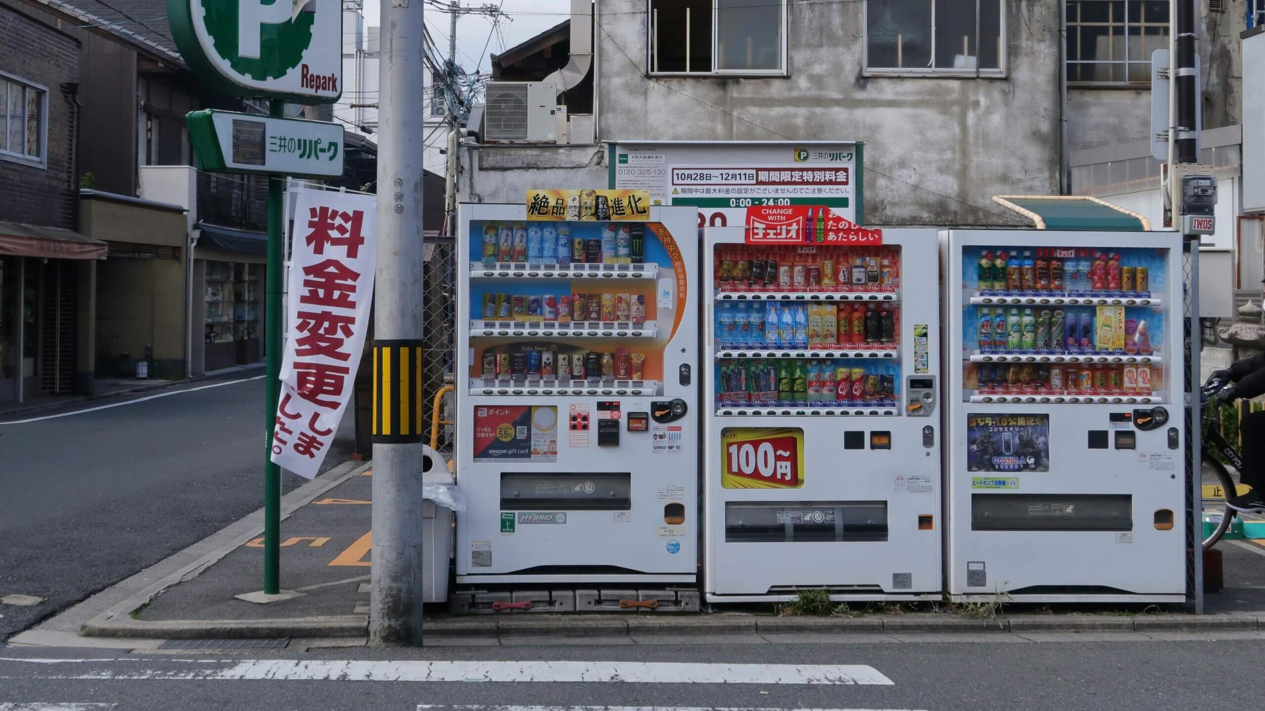 Three Japanese vending machines with various drinks on a city street corner near a Repark parking sign