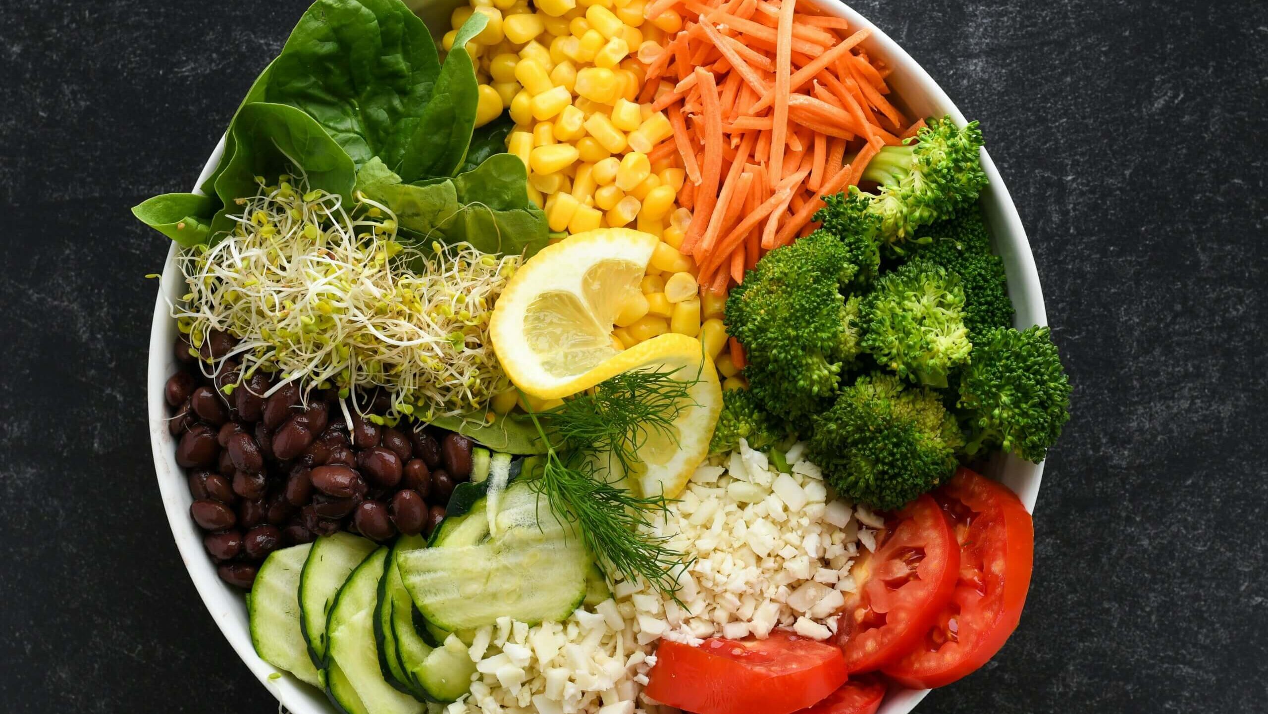 Bowl of fresh vegetables including spinach, corn, shredded carrots, broccoli, tomatoes, cucumber, black beans, sprouts, and lemon slices on dark surface