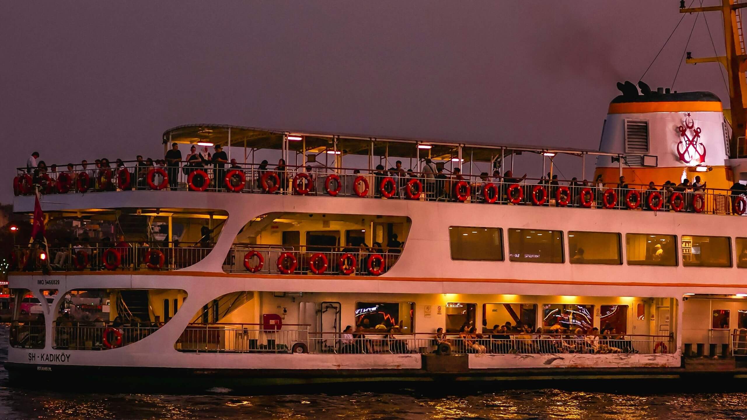 Passenger ferry SH-Kadıköy with people on board sailing at dusk in Istanbul.