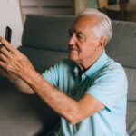 Elderly man in a light blue polo shirt sitting on a gray couch holding a smartphone up for a selfie or video call.