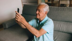 Elderly man in a light blue polo shirt sitting on a gray couch holding a smartphone up for a selfie or video call.