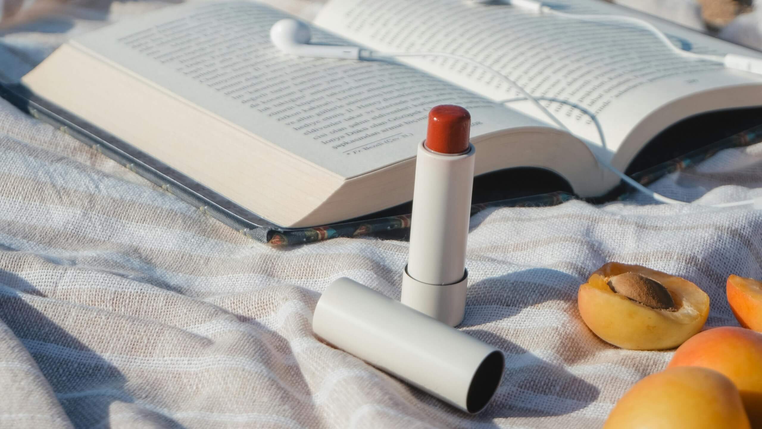 Open book with white earphones, apricot fruits, and a beige lipstick on a striped fabric surface outdoors.