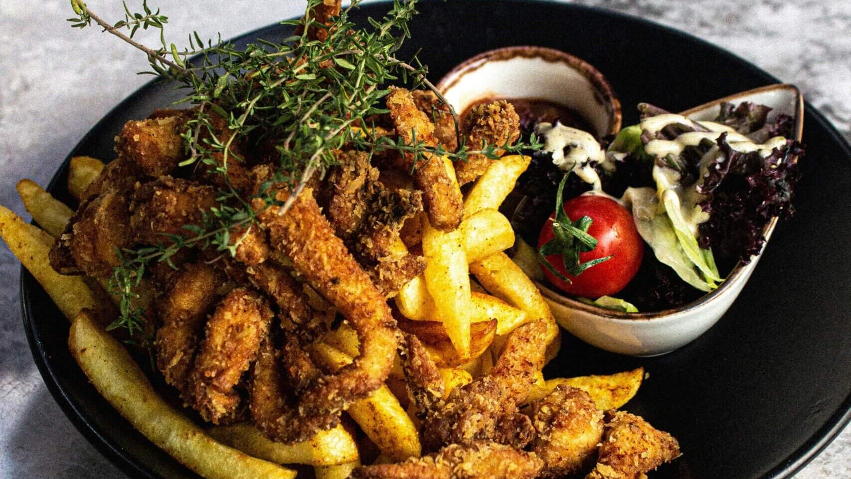 Fried calamari with French fries, fresh thyme, dipping sauce, and side salad with cherry tomato on a black plate