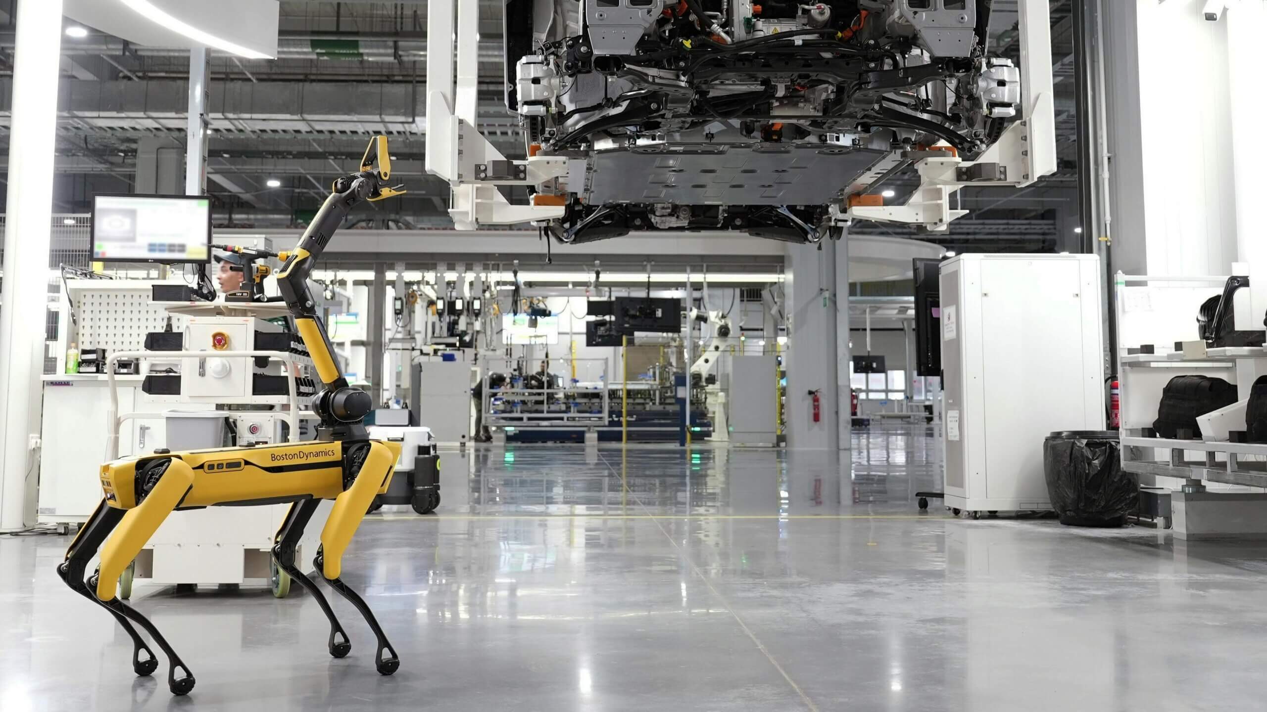Boston Dynamics robot dog with robotic arm in an industrial factory setting under a suspended vehicle chassis.