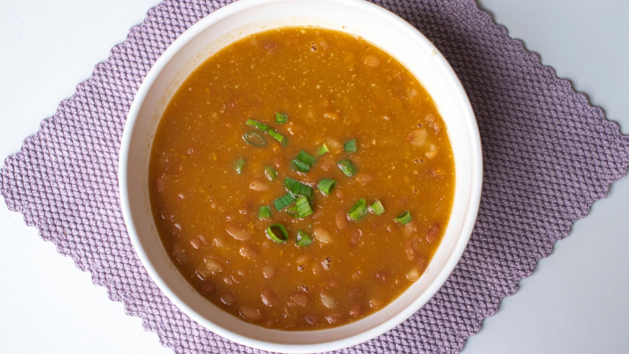 Bowl of bean soup garnished with chopped green onions on a purple textured placemat