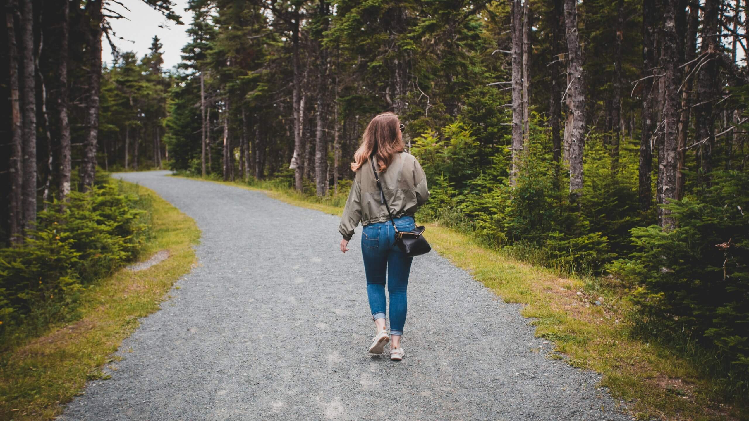 Woman in green jacket and jeans walking on a gravel path through a forested area