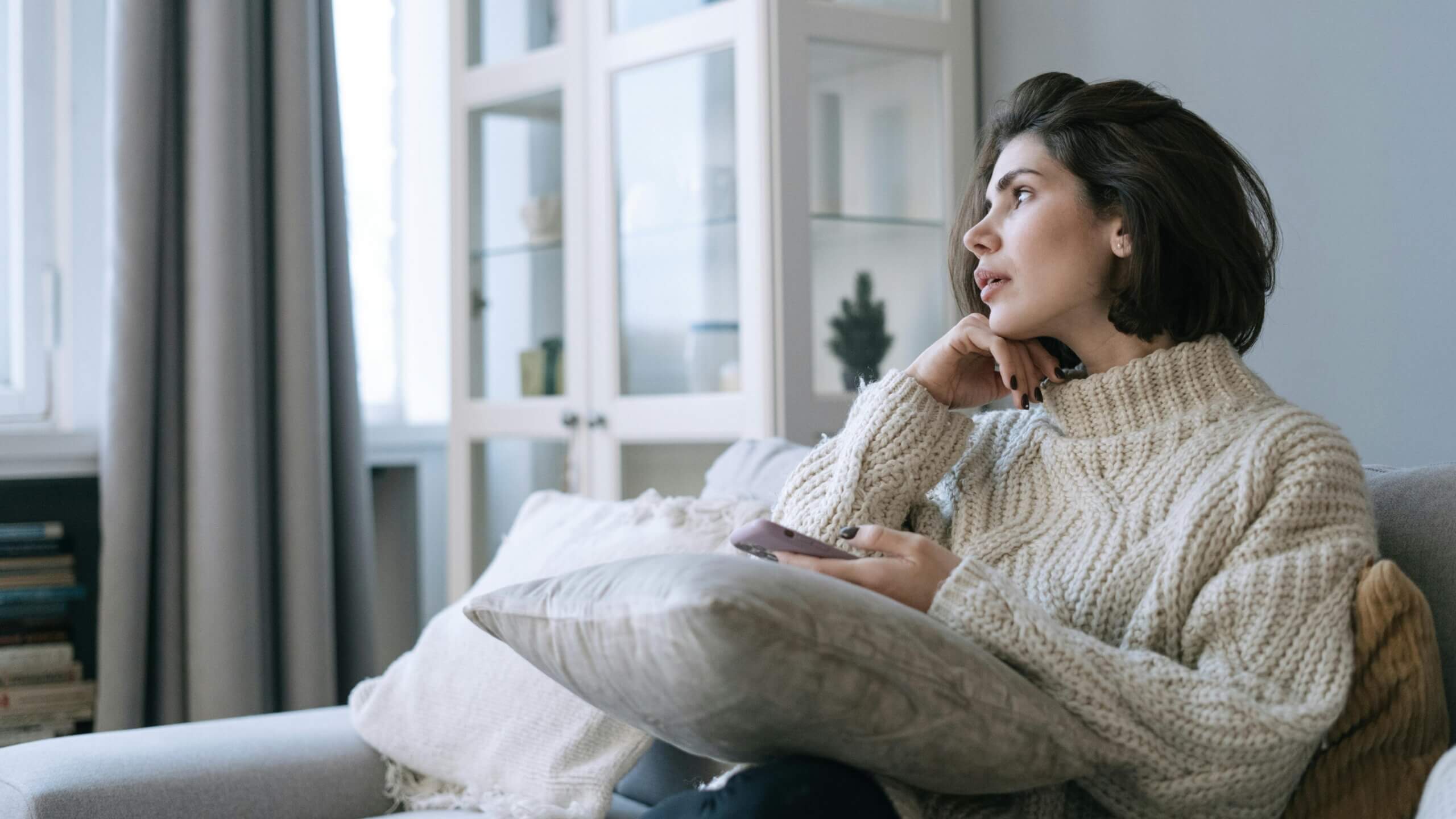 Woman in a cream knit sweater sitting on a couch holding a phone and looking thoughtfully out a window