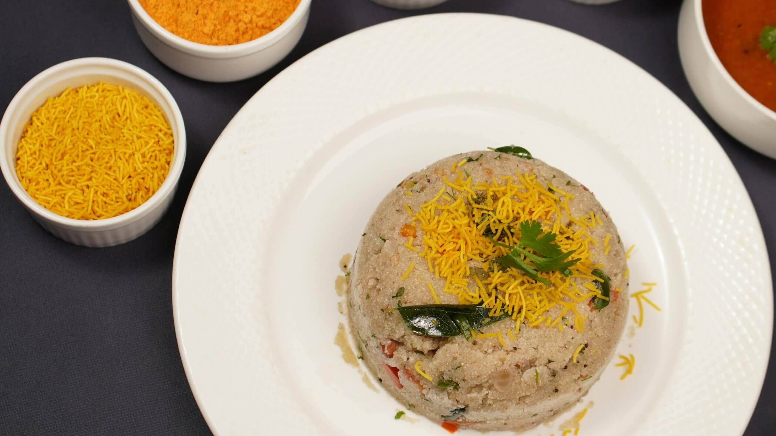 South Indian upma garnished with sev and curry leaves on a white plate with bowls of sev and chutney nearby