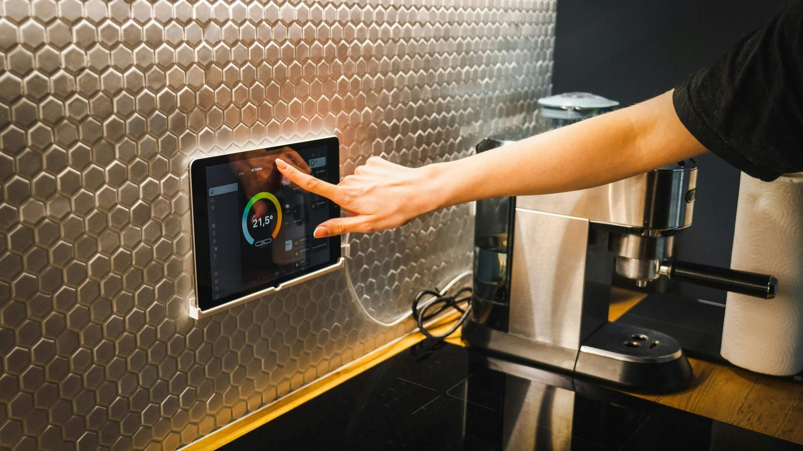 Hand adjusting a smart thermostat mounted on a hexagonal tiled kitchen wall next to a coffee machine.